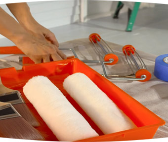 Person holding two paint rollers next to paint tray with two paint brushes in it