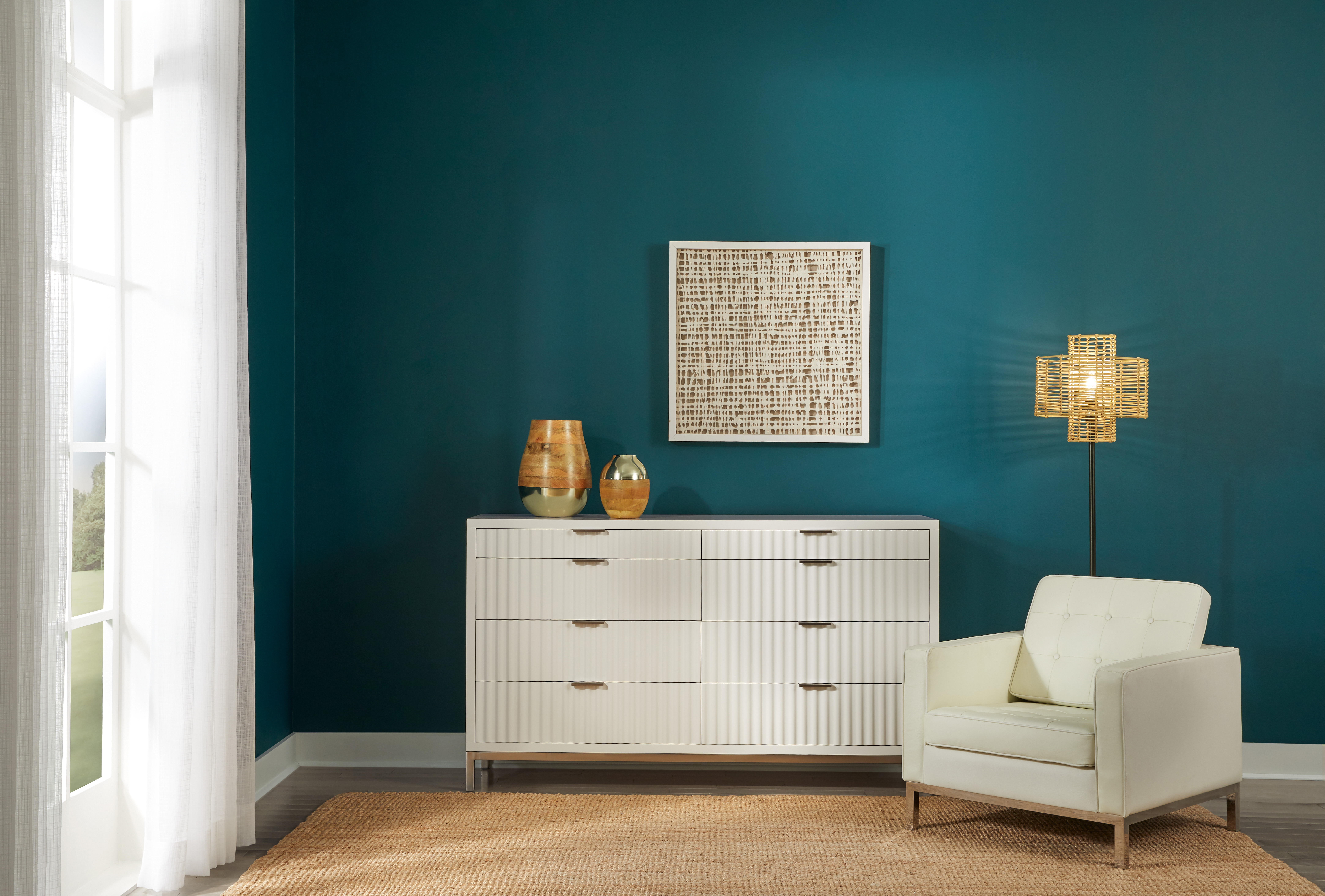 Un angle d'un côté d'une chambre, le mur est peint en sarcelle, teinte inspirée des profondeurs de l'océan et qui s'appelle Abîme d'océan. Il y a une commode de couleur claire et une chaise rembourrée devant le mur, ce qui crée un joli contraste entre le mur et les meubles.