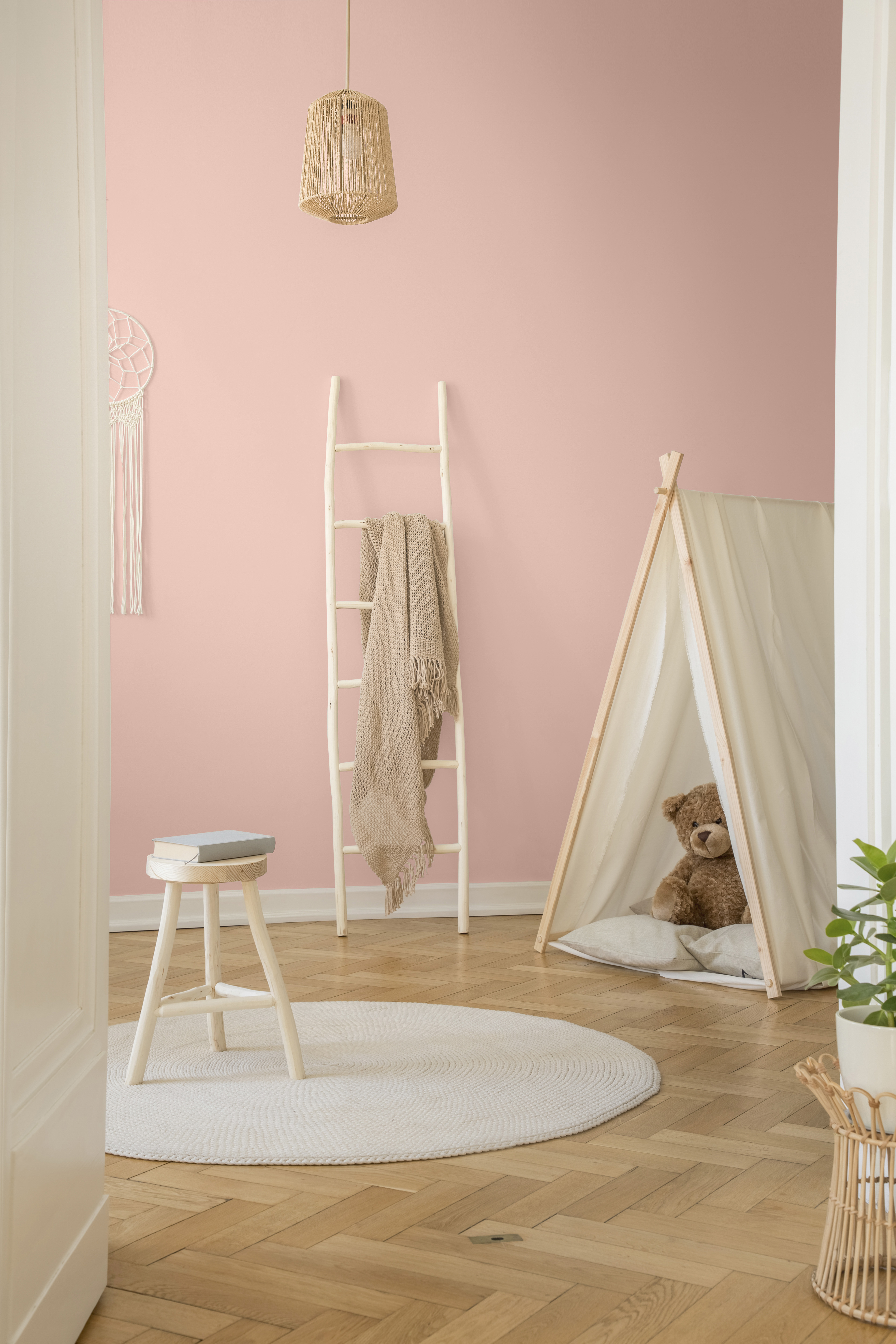 La vue verticale d'une chambre pour bébé blanche d'influence scandinave avec tente, ours en peluche, échelle en bois et couverture beige. Le mur du fond est peinturé en rose clair.