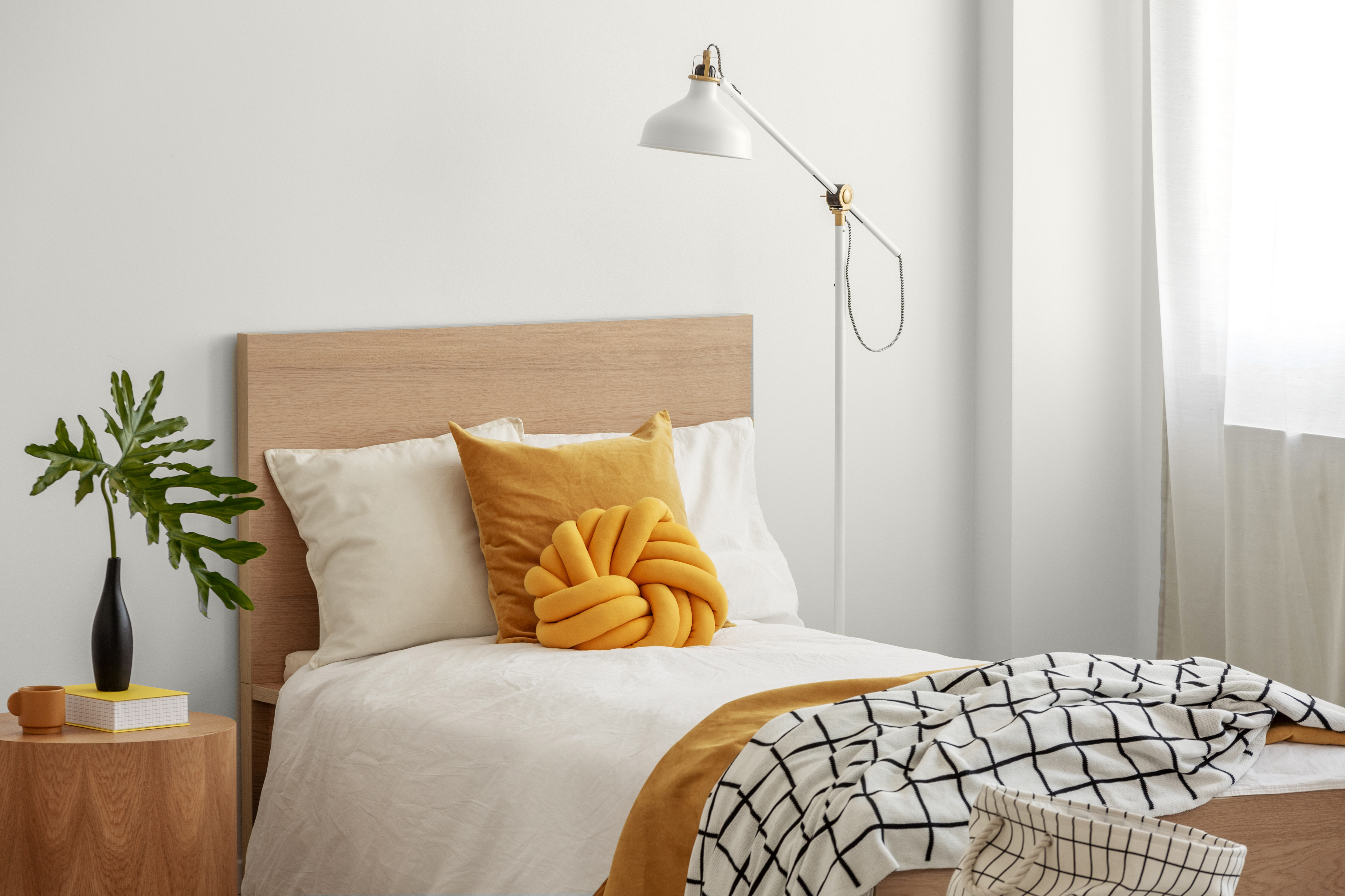 Des coussins jaunes et une couverture confortable sur un lit simple dans une élégante chambre d'hôtel, du feuillage vert dans un vase noir sur une table de chevet en bois.