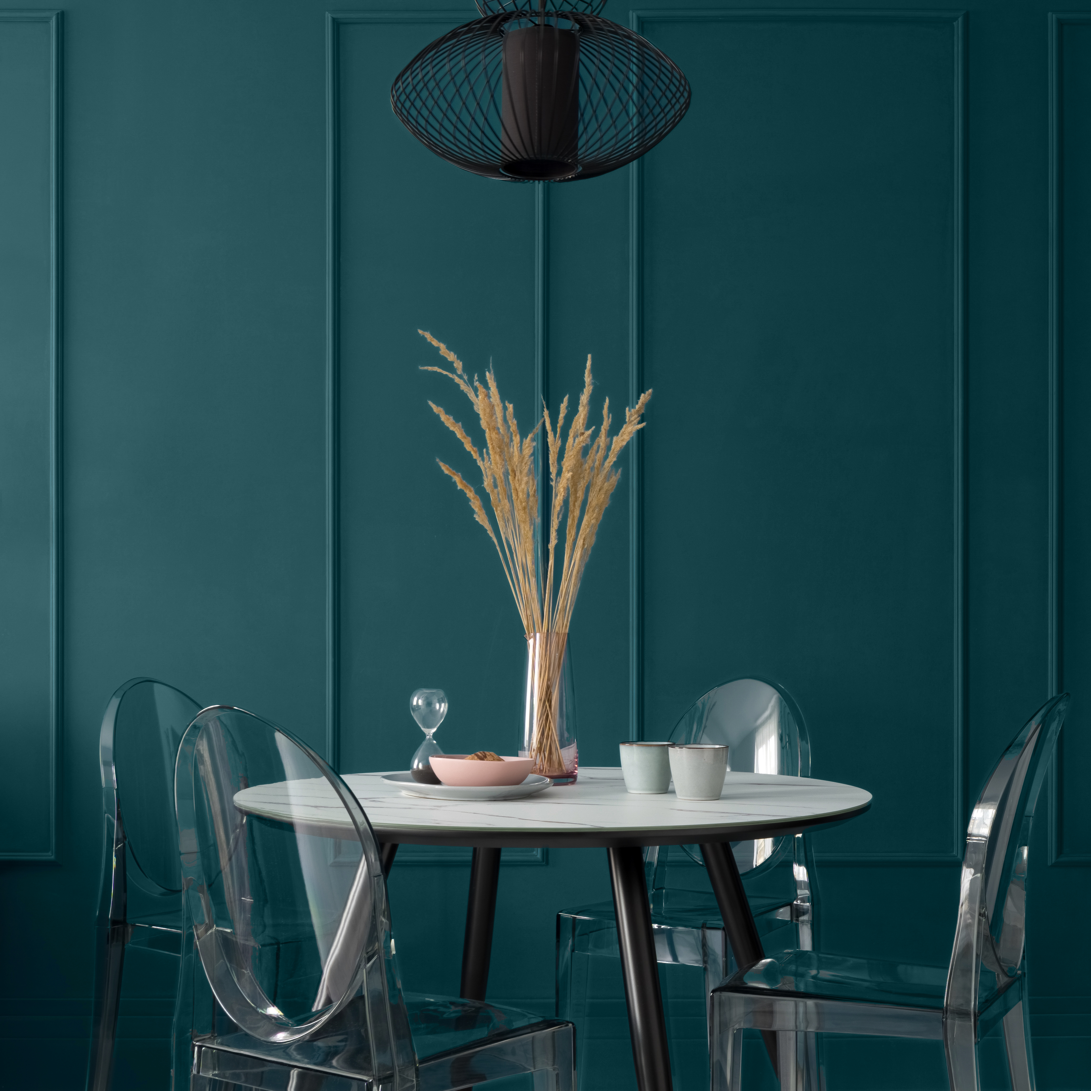 Une salle à manger distinguée avec un mur bleu-vert, une table moderne montée de pieds noirs avec un dessus de table de style marbre, de belles décorations de table, quatre chaises neuves en plastique sous une lampe suspendue noire. Le mur est peint dans une profonde teinte sarcelle appelée Abîme d'océan.
