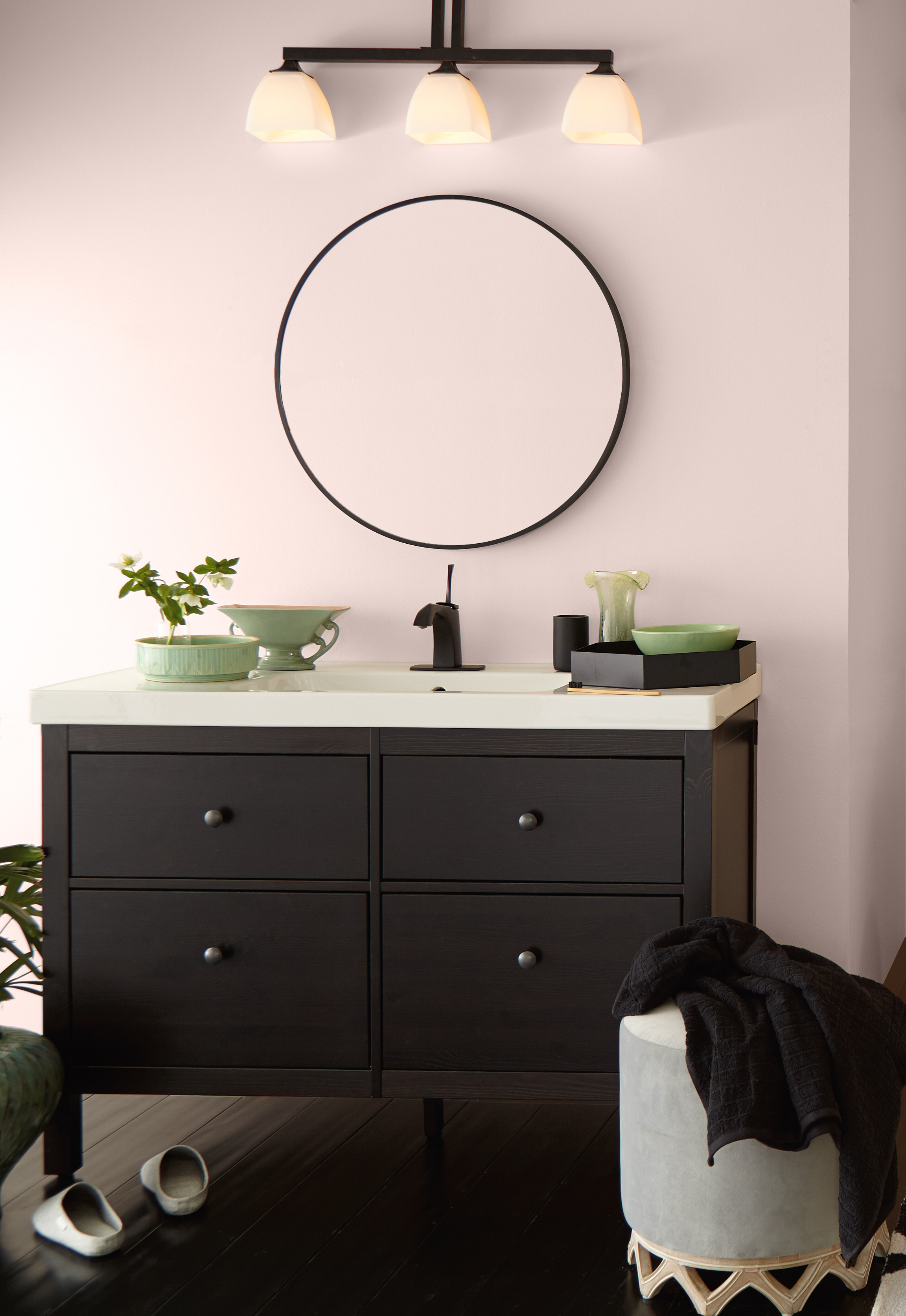 Une salle de bain met en valeur le meuble-lavabo décoré d'accents vert sauge. Un miroir circulaire et des murs peints en Baiser volé, un ton délicat de rose clair.
