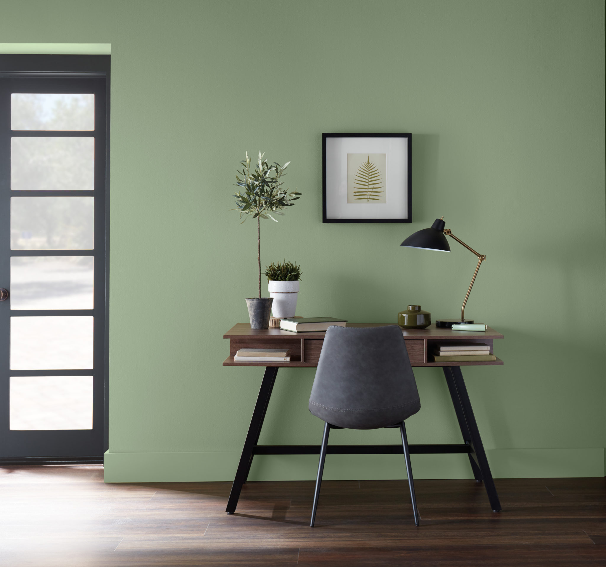 Un coin bureau décontracté dans un salon avec un pupitre  moderne et une chaise confortable. Pour compléter le look, on retrouve quelques éléments décoratifs de bureau. La couleur du mur est un vert moyen appelé Arbre de lauriers.