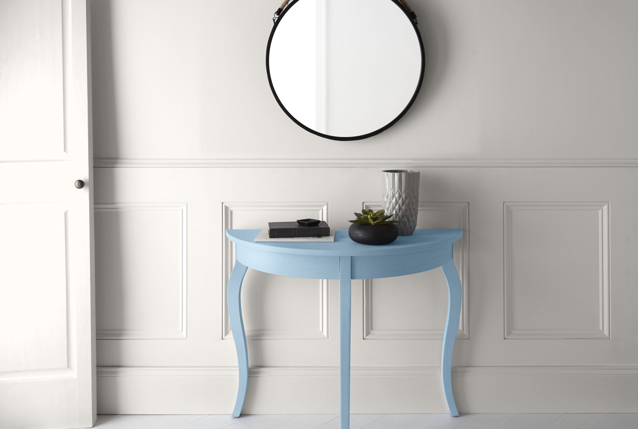 Un couloir tout blanc avec une petite console bleue. On y voit un simple miroir rond et des objets de décoration sur le dessus de la table.