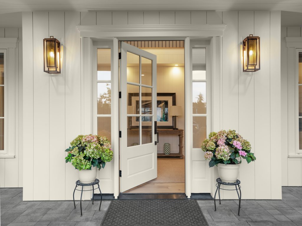 Une entrée de maison entièrement blanche avec une porte ouverte, quelques plantes décoratives soigneusement sélectionnées et des luminaires simples créent un sentiment de paix et de grâce. Un excellent exemple de "moins, c'est plus".