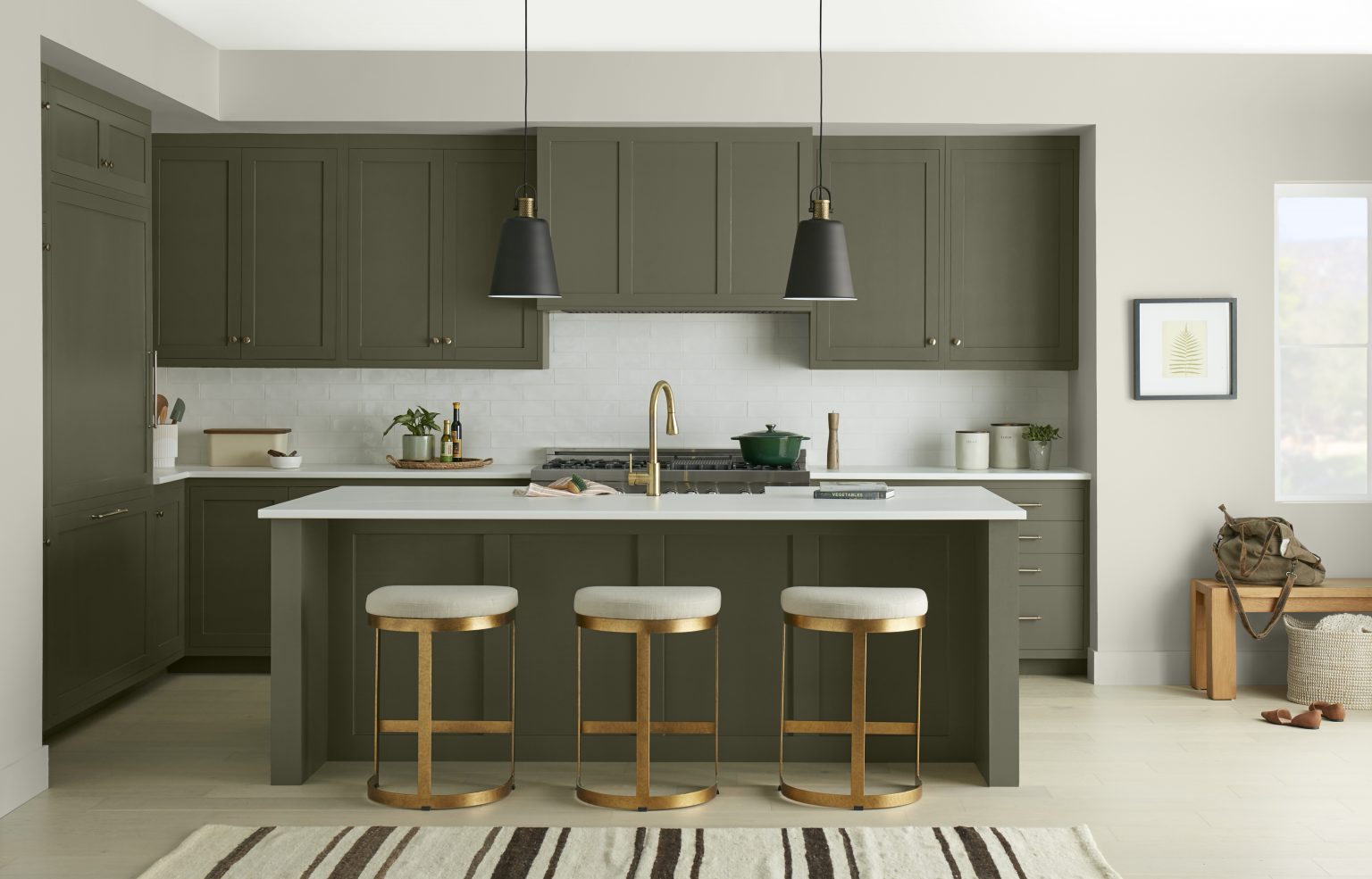 Une cuisine moderne aux armoires et îlot peints en vert olive foncé et une quincaillerie et tabourets dorés 