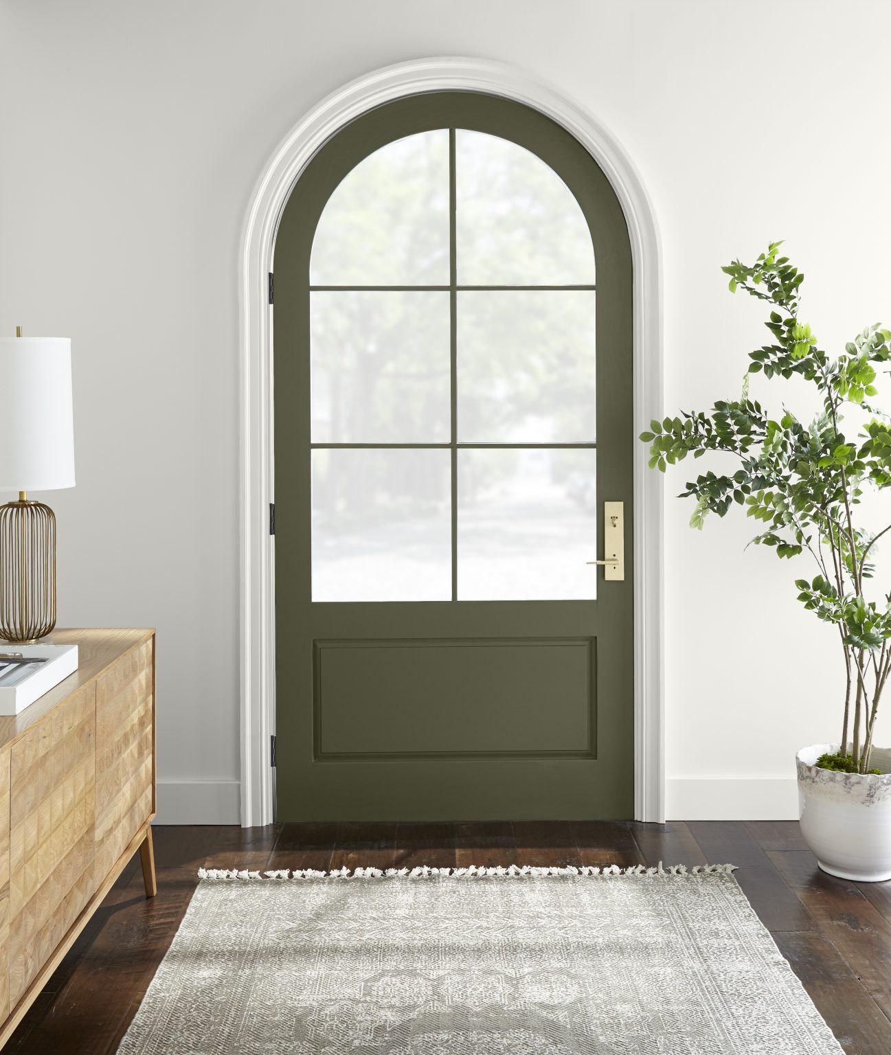Une entrée peinte en blanc avec une porte voûtée verte, une table console en bois, un tapis neutre et une plante