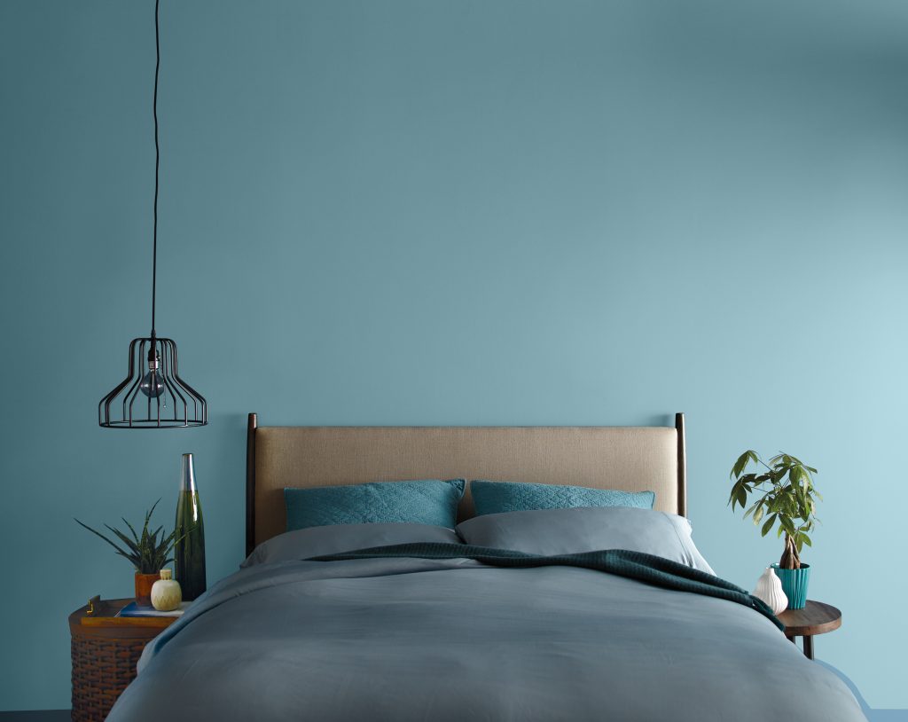 Une chambre à coucher avec le mur de tête de lit peint en bleu atténué, décorée d’un cadre de lit neutre, de draps et oreillers bleus et de tables de nuit