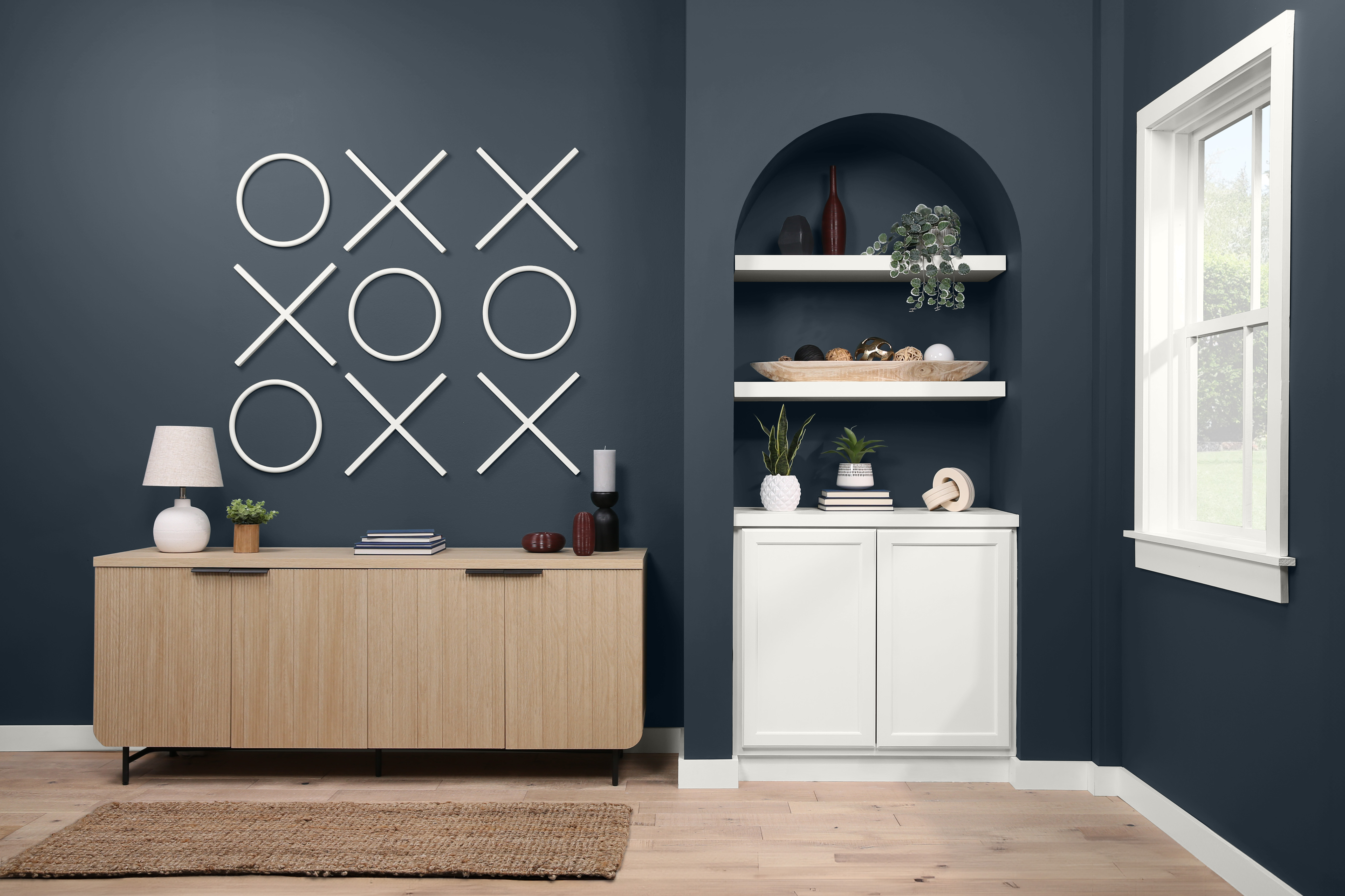 L'intérieur d'un élégant bureau à domicile au décor ludique, avec un grand tic-tac-toe en guise d'art mural. Un buffet moderne est orné d'une lampe blanche et une étagère encastrée avec un éventail d'objets décoratifs et de verdure.  La pièce bleu foncé est dotée d'une fenêtre qui laisse passer beaucoup de lumière, ce qui renforce l'atmosphère sereine et moderne.