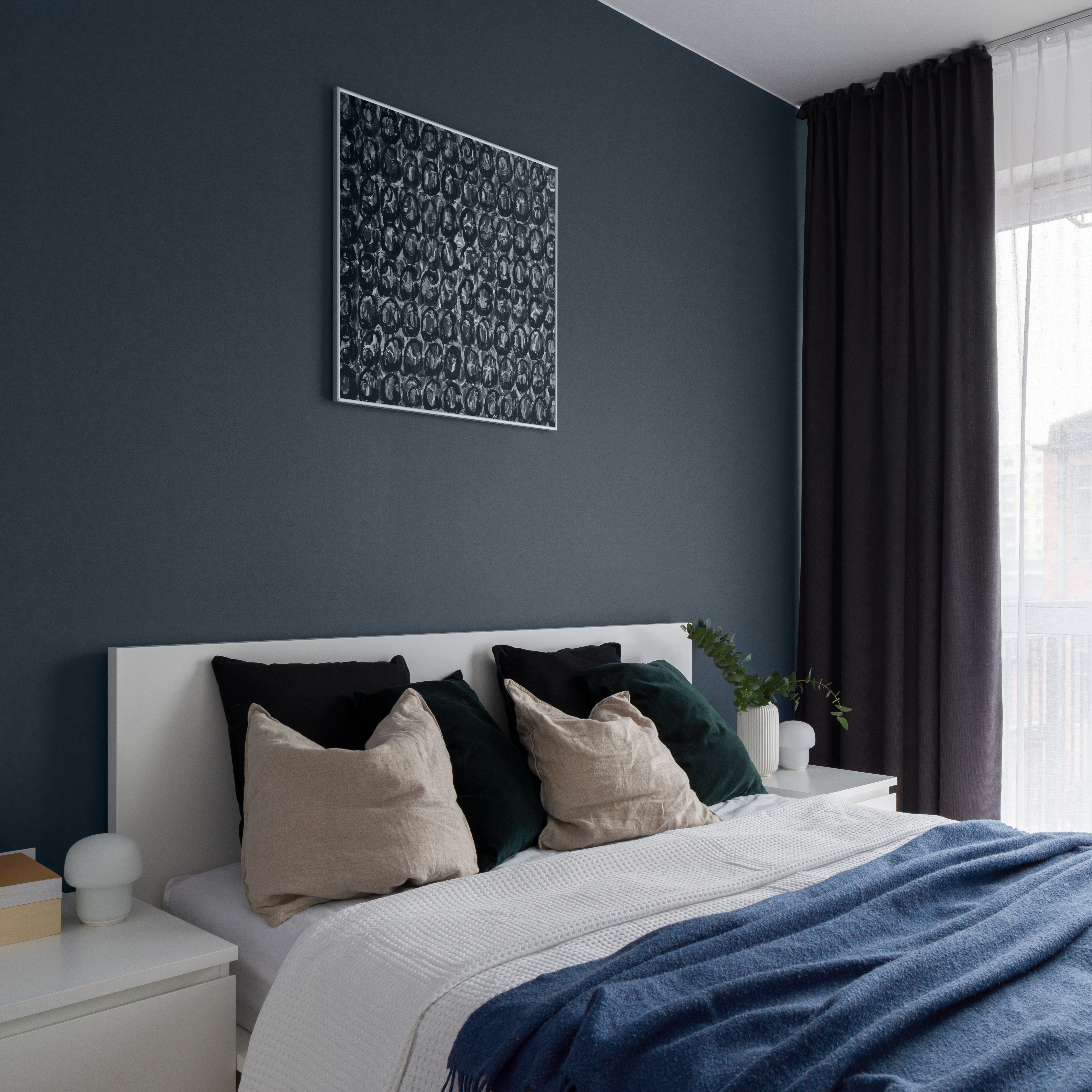 Une chambre à coucher contemporaine au design minimaliste, avec un lit blanc, de la literie fraîche et une couverture bleue. La pièce est agrémentée d'un mur d’accent bleu foncé peint avec une couleur BEHR appelée Bleu minuit. Les rideaux gris foncé sont ouverts pour laisser entrer la lumière naturelle, complétant ainsi la palette de couleurs sereine et coordonnée de la chambre.

