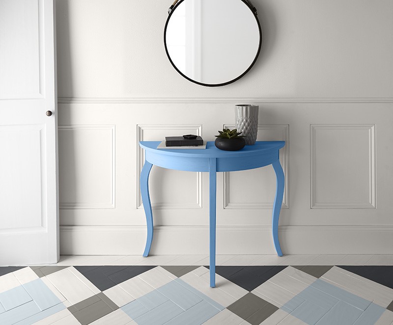 Un couloir avec des murs blancs, des garnitures blanches et une porte blanche. Une table bleue en demi-lune est placée contre le mur sous un miroir rond au cadre noir, ajoutant une touche de couleur à ce décor. Le plancher présente un motif géométrique composé de carreaux pâles et foncés, ce qui contribue à l'ambiance moderne de la pièce.
