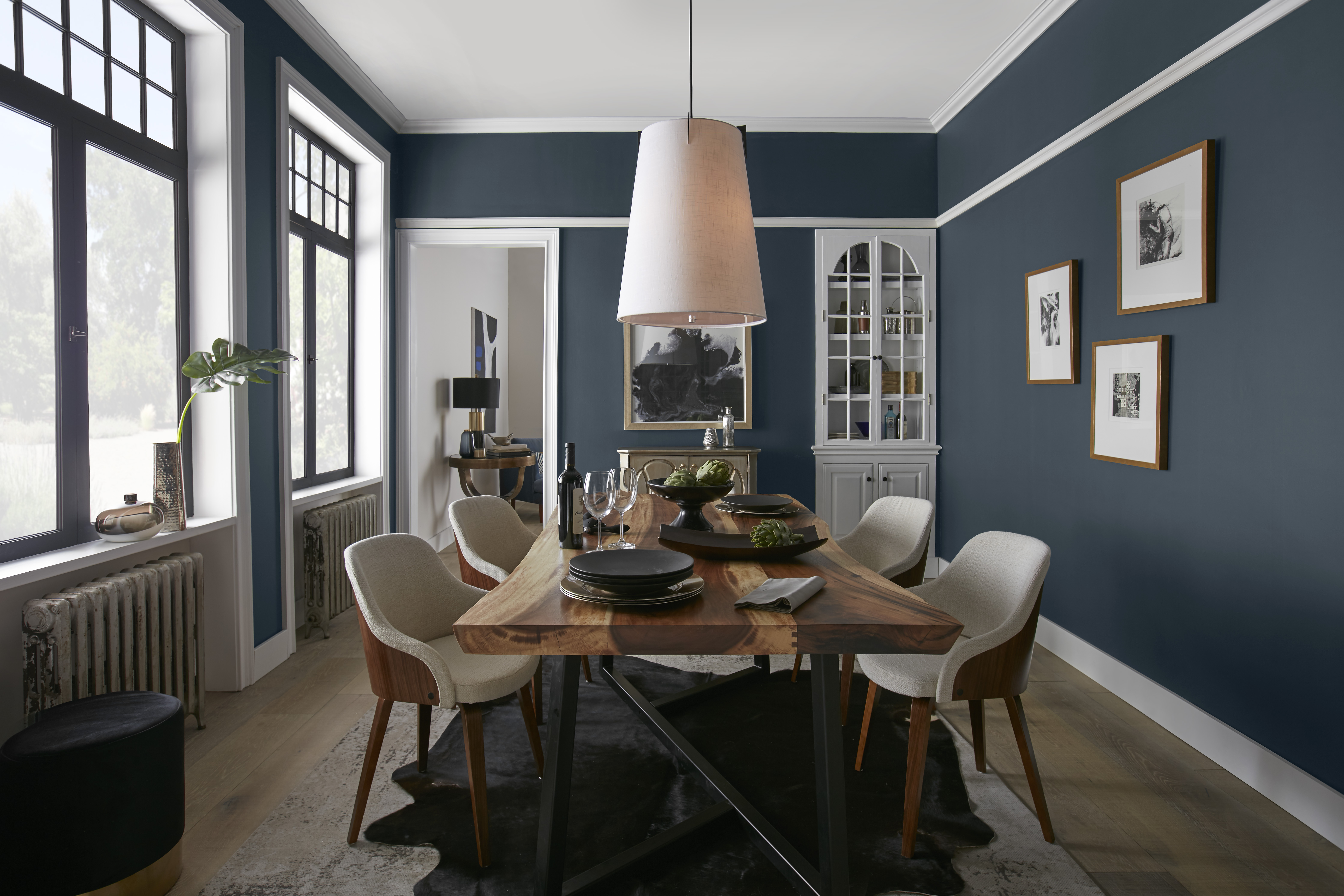 Une salle à manger chic avec une longue table en bois entourée de chaises rembourrées beiges, aux murs bleu foncé et garnitures blanches. Une lampe suspendue à l'abat-jour texturé jette une lumière douce sur la table, qui est soigneusement dressée avec des assiettes et des verres, et ornée d'une plante verte en guise de centre de table.

