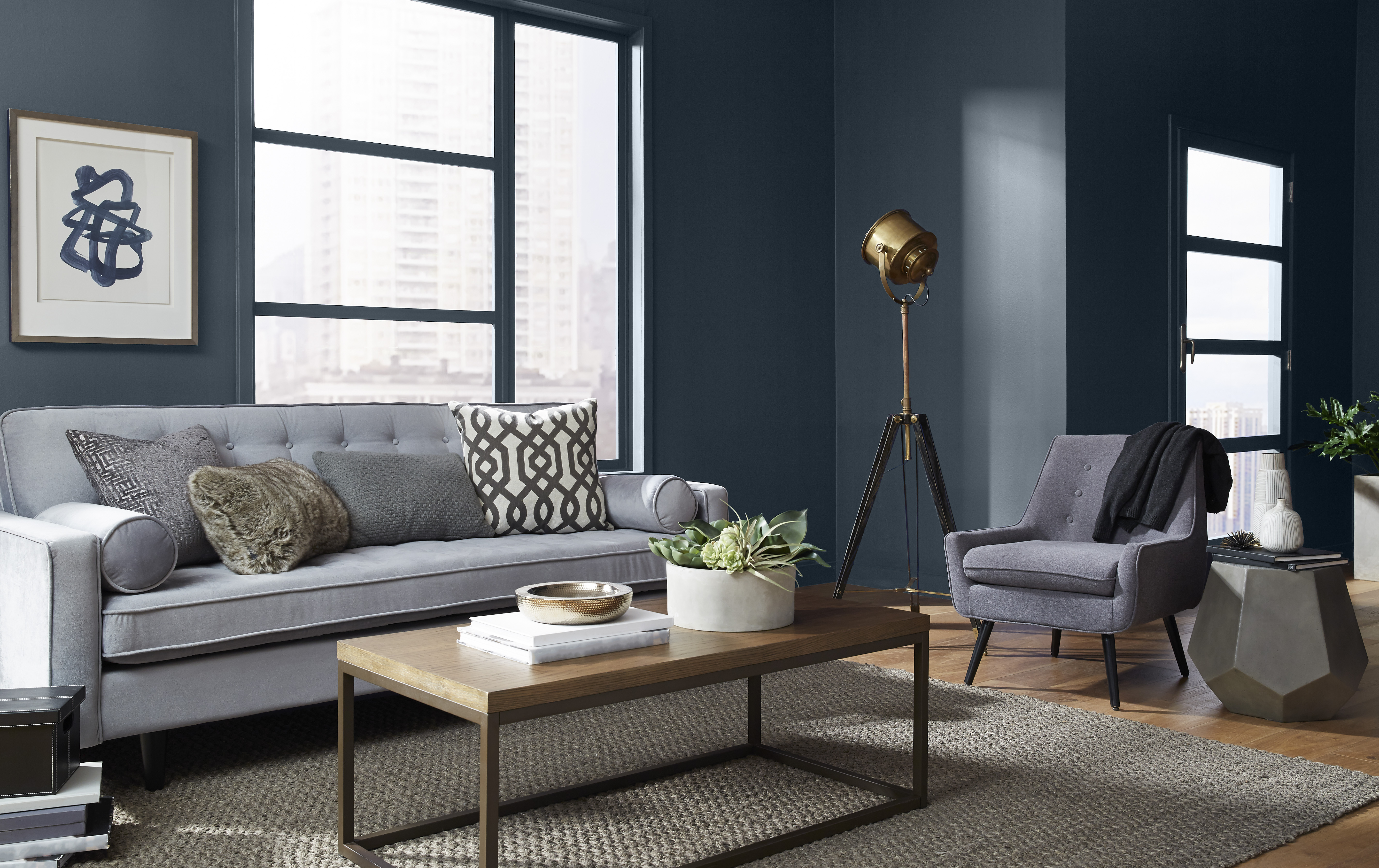 Un salon moderne et confortable avec un sofa gris rembourré, orné de coussins à motifs, et un fauteuil assorti à côté d'une table d'appoint géométrique unique. Un lampadaire en laiton ajoute de la chaleur au coin, tandis que les murs d'un bleu profond, rehaussés de garnitures blanches, encadrent de grandes fenêtres qui révèlent une vue sur le paysage urbain.

