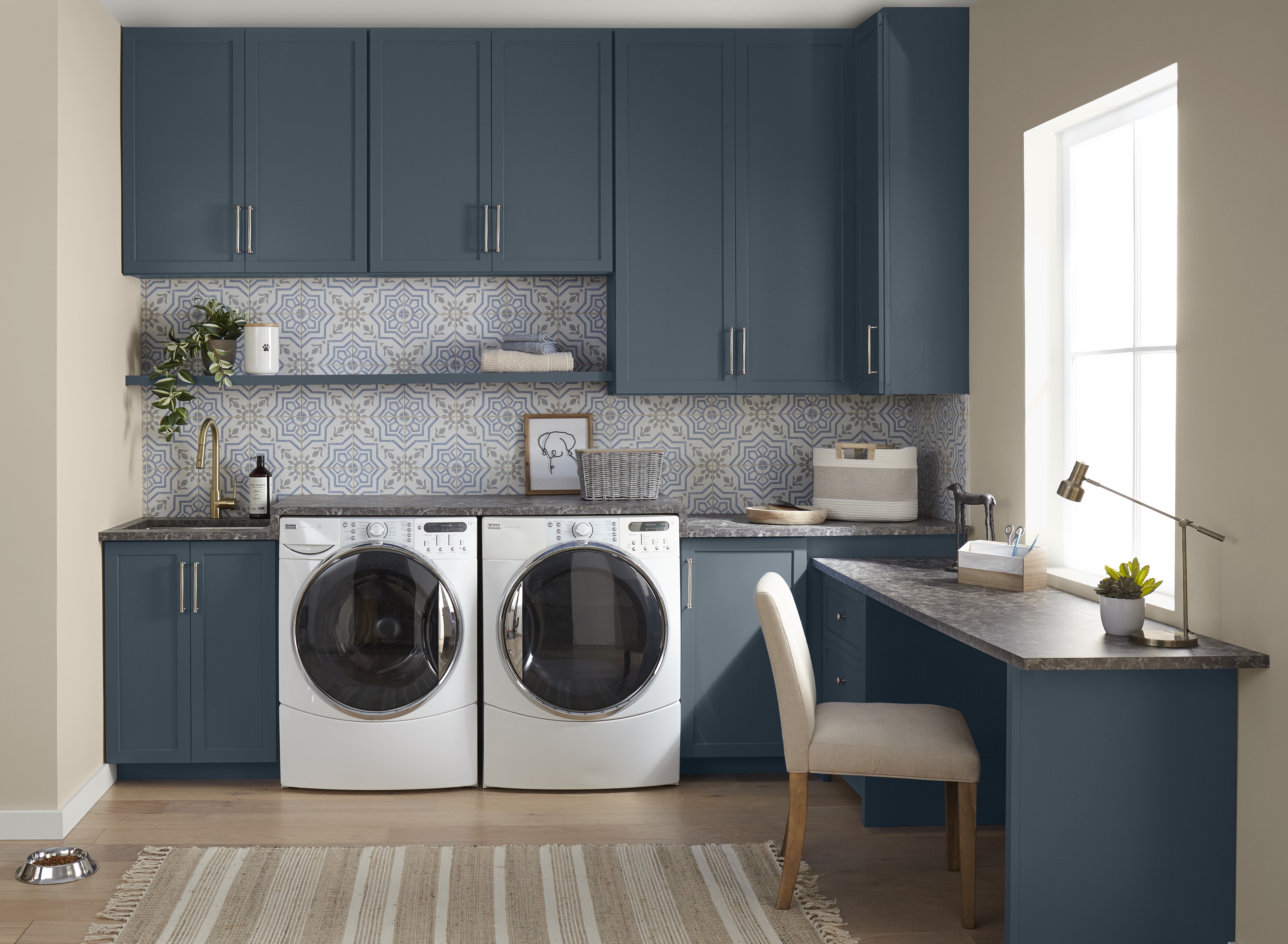 Une buanderie/bureau à domicile avec des armoires bleu foncé qui contrastent magnifiquement avec des murs beiges et un dosseret en carreaux à motifs gris clair et bleus. Les garnitures sont en Ours polaire, un blanc éclatant, ce qui ajoute à l'aspect frais et propre de l'ensemble.