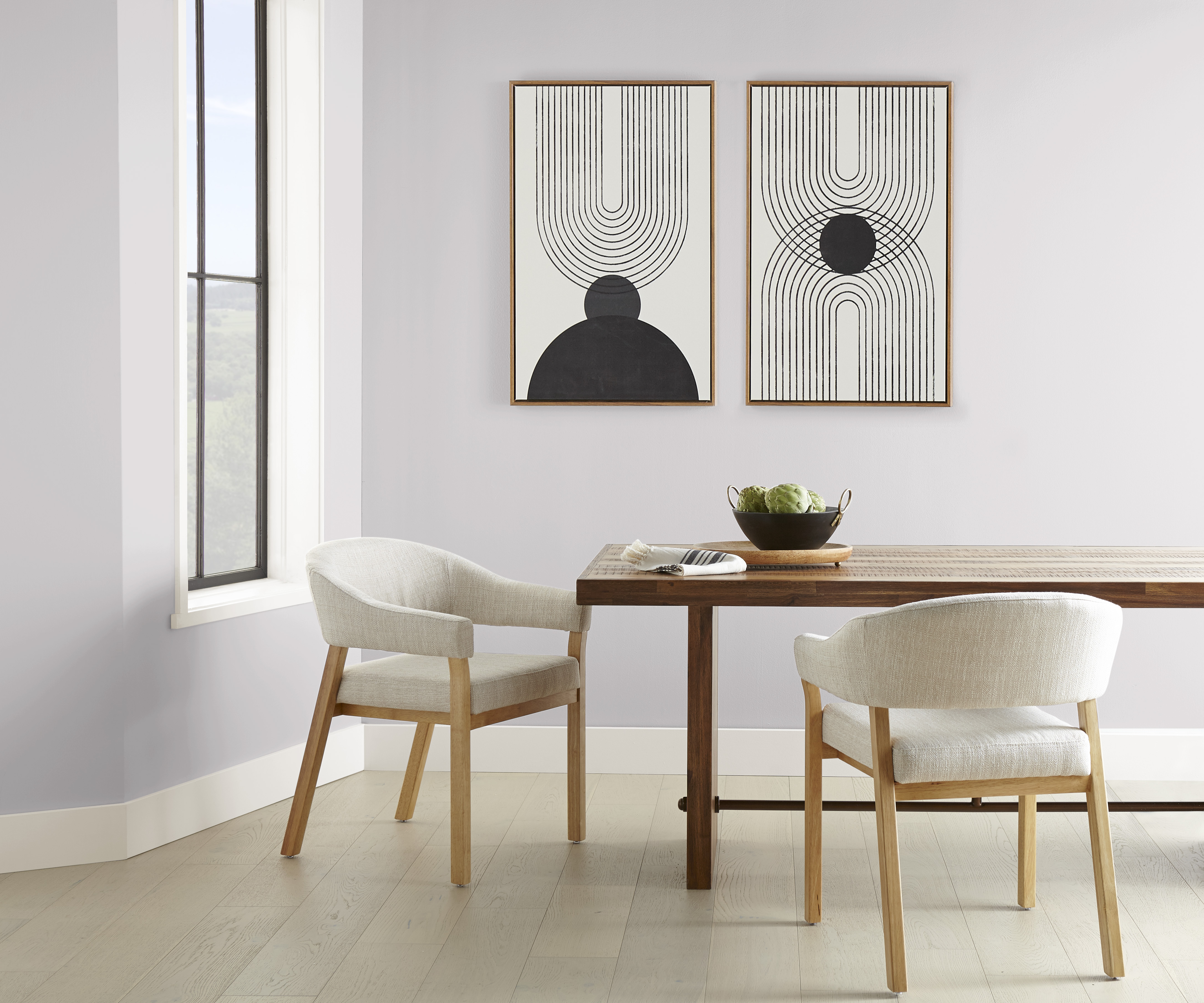 Cette image montre une salle à manger moderne au design minimaliste avec une table en bois flanquée de deux chaises rembourrées de couleur claire. Sur la table, un bol noir contient des artichauts. Le mur derrière la table affiche deux œuvres d’art abstraites constituées de motifs géométriques en noir et blanc. Une grande fenêtre sur le côté gauche laisse pénétrer la lumière du jour, mettant en valeur le parquet en bois clair et les murs peints en Cristal de roche.