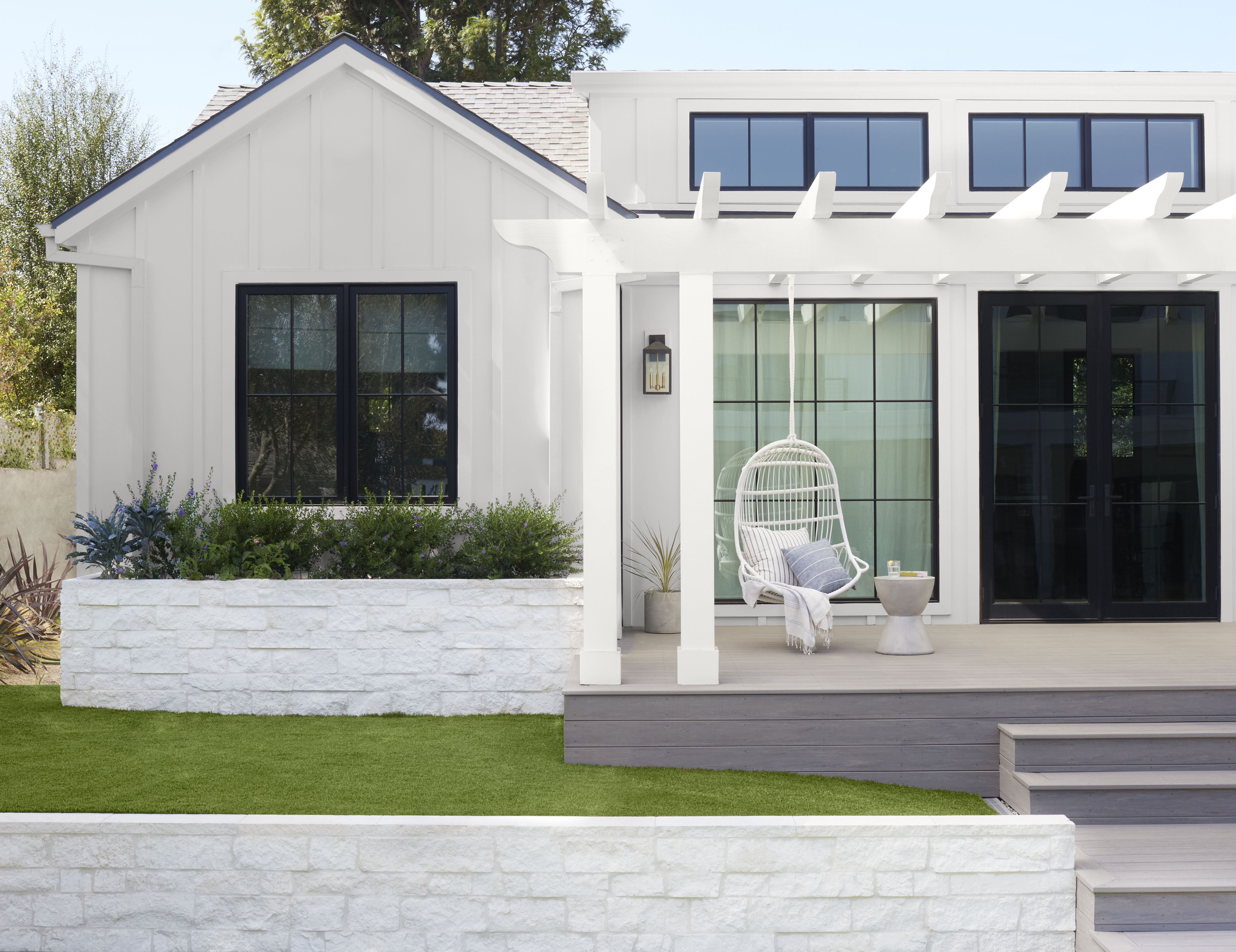 Cette image montre une maison de couleur claire avec un porche d’entrée doté d’une balançoire suspendue à une pergola, créant un espace extérieur invitant et confortable.