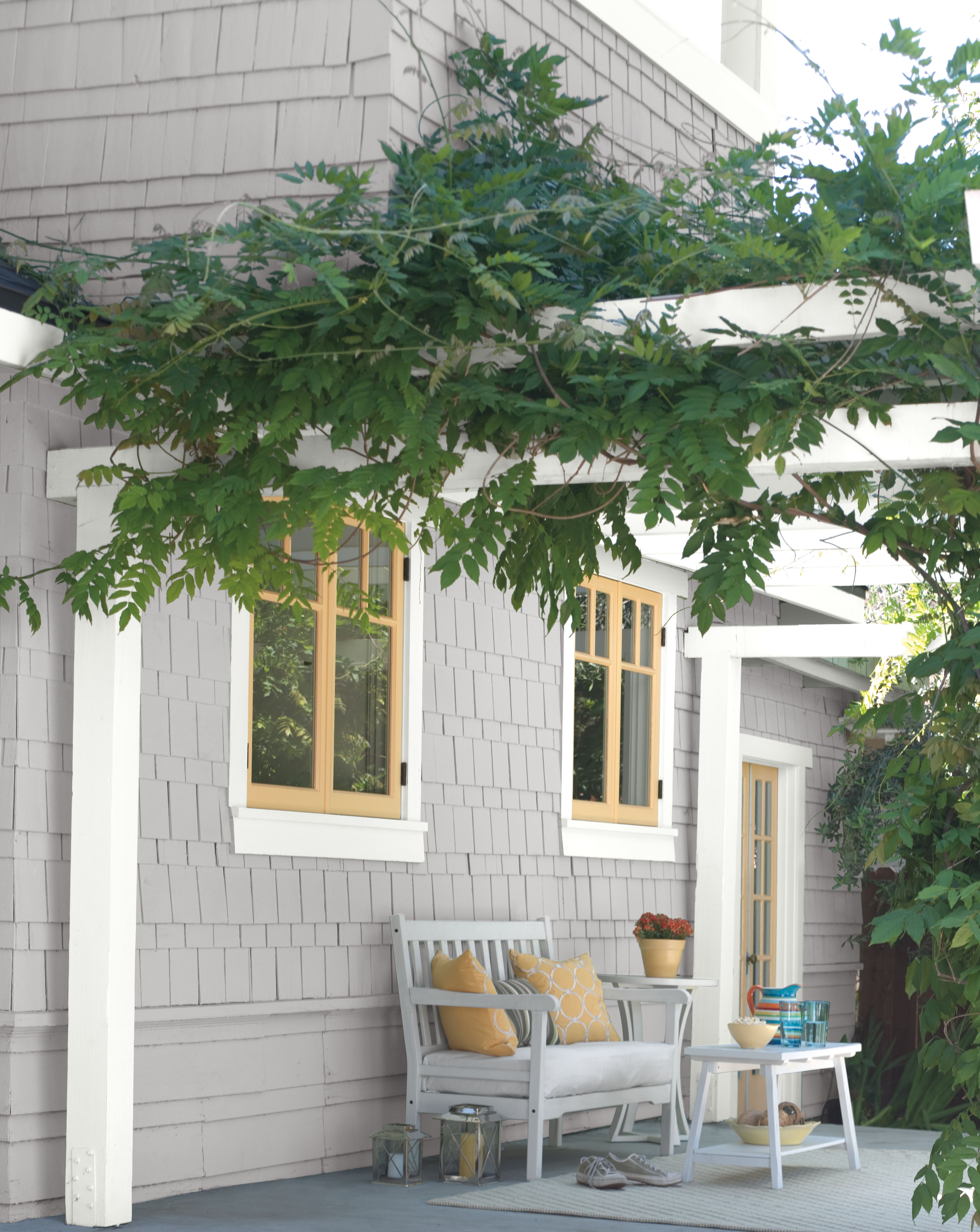 Cette image montre le porche d’un charmant cottage avec des murs extérieurs peints en gris et des garnitures blanches, le tout joyeusement accentué de fenêtres et d’une porte jaunes, créant une oasis lumineuse et invitante.