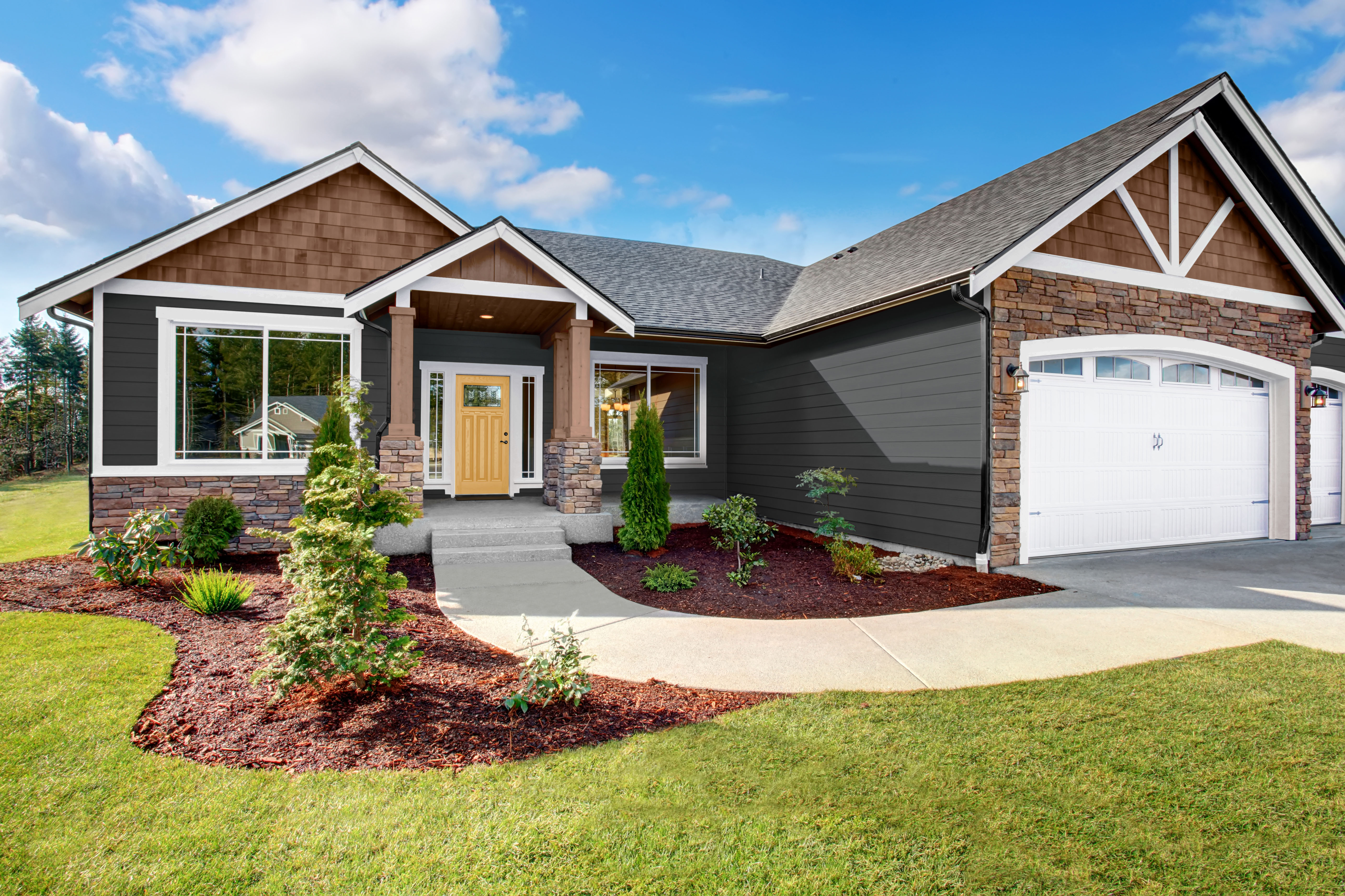 Cette image montre une maison artisanale de style ranch. Elle est peinte en gris anthracite foncé avec des garnitures d’un blanc éclatant, le tout accentué de bardeaux en bois de couleur naturelle et d’un revêtement en pierre. La porte d’entrée jaune vif apporte une joyeuse touche de contraste, conférant chaleur et personnalité à l’extérieur classique.