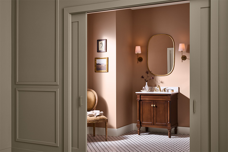 Cette image montre une élégante salle de bain avec des murs rose pâle, un meuble-lavabo en bois avec un dessus blanc et un miroir ovale à cadre doré. Deux appliques murales avec abat-jours de couleur pêche flanquent le miroir, et une chaise rembourrée, sur laquelle reposent des serviettes pliées, se dresse contre le mur latéral sous deux petits tableaux encadrés.