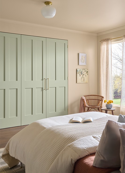 Cette image montre une chambre à coucher confortable et décontractée, avec un lit recouvert d’une couette beige sur laquelle repose un livre ouvert. La lumière naturelle pénètre à flots par une grande fenêtre habillée de rideaux diaphanes. Dans le coin de la pièce, juste à côté de la fenêtre, on aperçoit un fauteuil en osier et une petite table ronde avec un vase de fleurs jaunes. Un placard à trois portes peintes en vert pâle et dotées de poignées dorées occupe le mur du fond, où deux œuvres d’art suspendues à droite saluent les occupants.