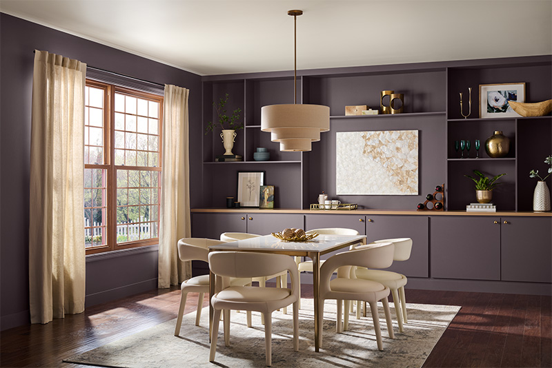Cette image montre une salle à manger éclectique avec des murs violet foncé et une immense fenêtre avec des rideaux beiges tirés sur les côtés. La pièce est meublée d’une table à manger blanche entourée de chaises blanc cassé sur un tapis de couleur claire. La table est éclairée par un plafonnier rond à gradins de couleur beige. Au fond de la pièce, le mur est doté d’étagères et d’armoires encastrées et ornées de divers objets, incluant des vases, des livres, des plantes et des œuvres d’art.