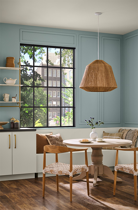 Cette image montre un coin repas lumineux avec des murs bleu-vert pâle et une grande fenêtre à carreaux dont le cadre et les grilles sont peints en noir. Une table ronde sur piédestal en bois est entourée de chaises à assise tressée et d’une banquette encastrée où sont disposés des coussins aux tons neutres.