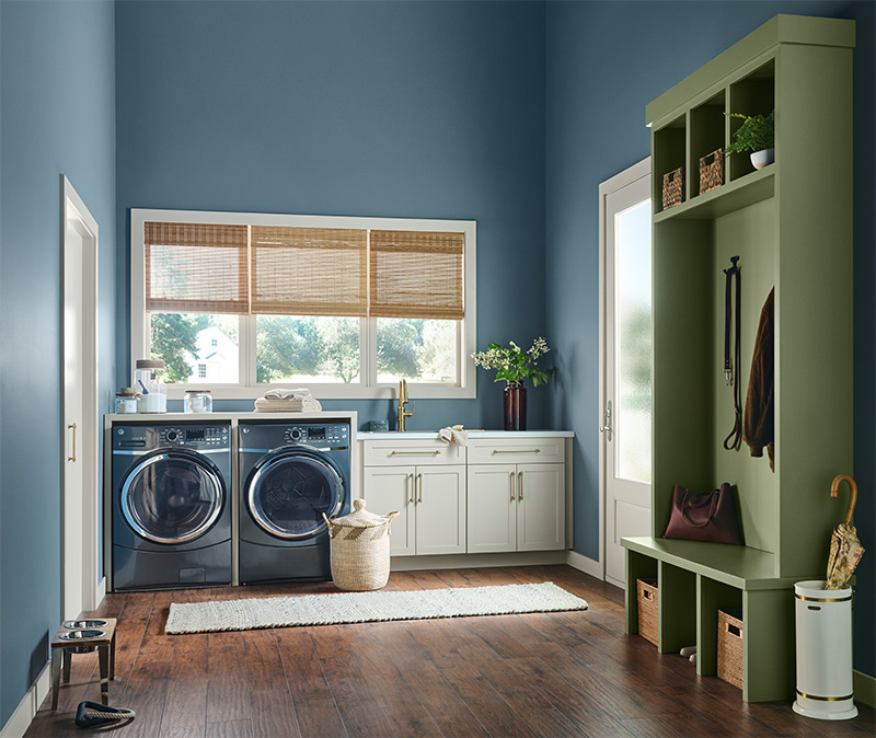 Cette image montre une salle de lavage avec des murs bleus et un plancher en bois. Elle comprend une laveuse et une sécheuse à chargement frontal, un plan de travail blanc avec des armoires en dessous et une série de trois fenêtres côte à côte surmontées de stores en bambou. À droite, un grand meuble de rangement peint en vert tient lieu de vestiaire d’entrée avec un banc, des crochets et des paniers de rangement en osier. Des articles ménagers, tels que des plantes, des bocaux et des produits d’entretien, complètent le décor de la pièce tout en lui donnant un air agréablement fonctionnel.