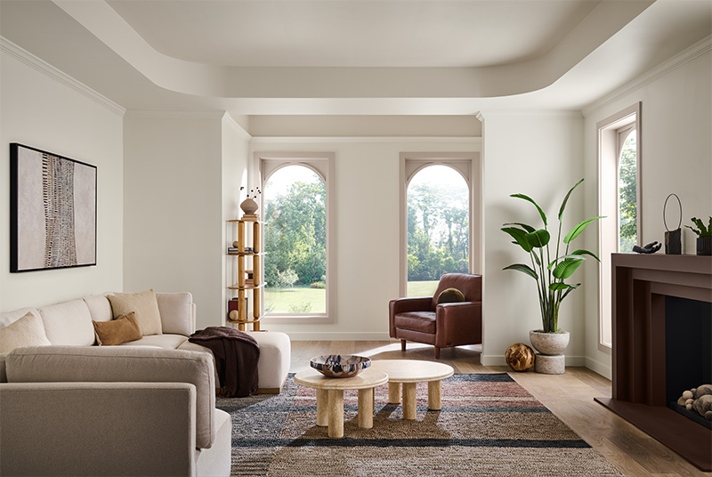 Cette image montre un salon moderne avec deux grandes fenêtres cintrées situées côte à côte et donnant sur le jardin. La pièce comprend un sofa beige avec des coussins, un fauteuil en cuir marron et une table basse en bois blond en plein centre. Une grande plante en pot ajoute de la vitalité au décor, de même qu’une plante plus petite sur le manteau de la cheminée. Une tour-étagère en bois ornée d’objets décoratifs est placée près d’une fenêtre, et un tableau d’art abstrait est accroché au-dessus du sofa. Les murs de la pièce sont blancs, et le plancher est couvert d’un tapis à motifs aux tons fauves.