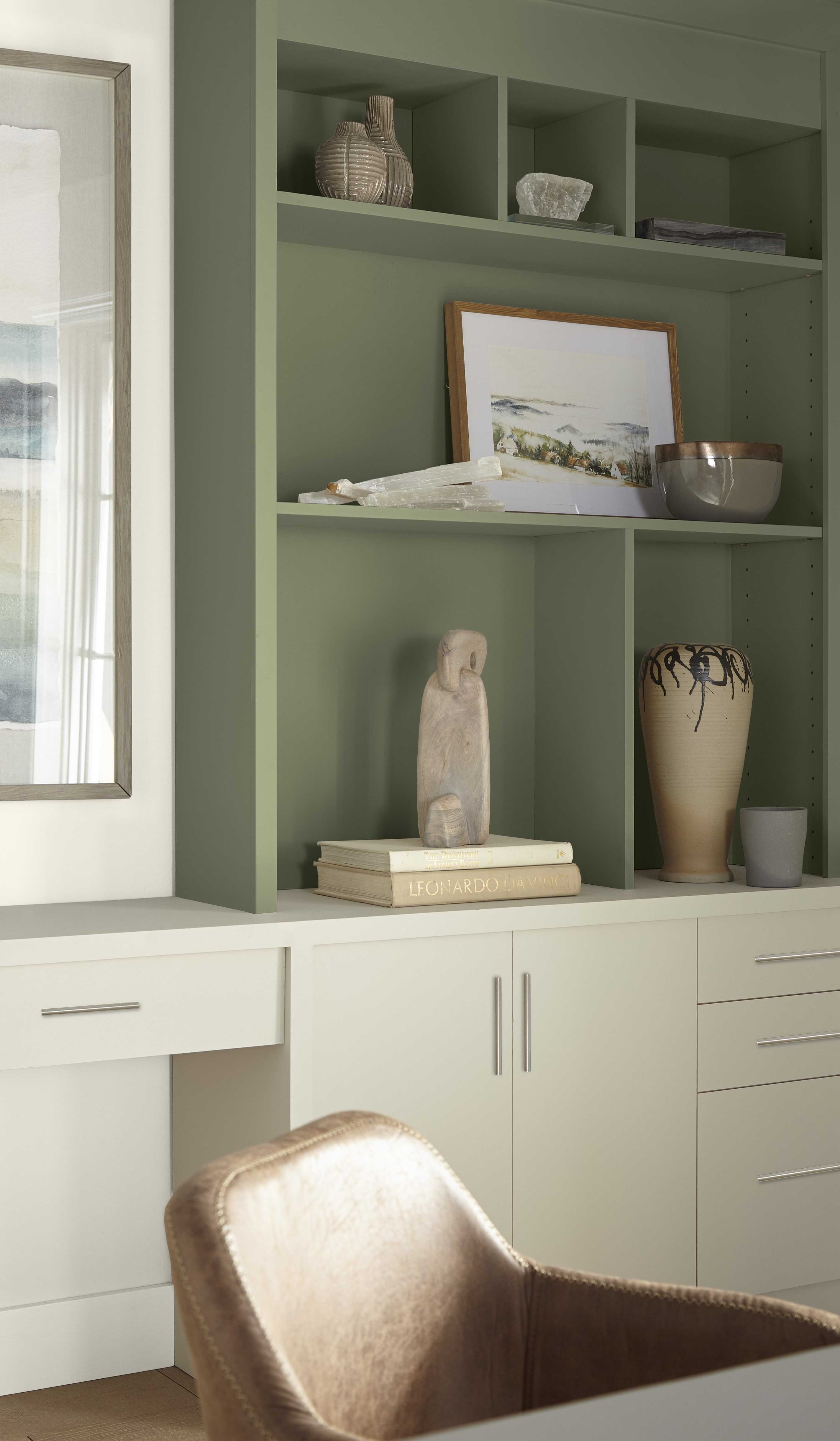 Cette image montre une pièce qui sert de bureau à domicile, avec une étagère encastrée peinte en vert sauge, des armoires blanches en dessous et un fauteuil en cuir brun clair.