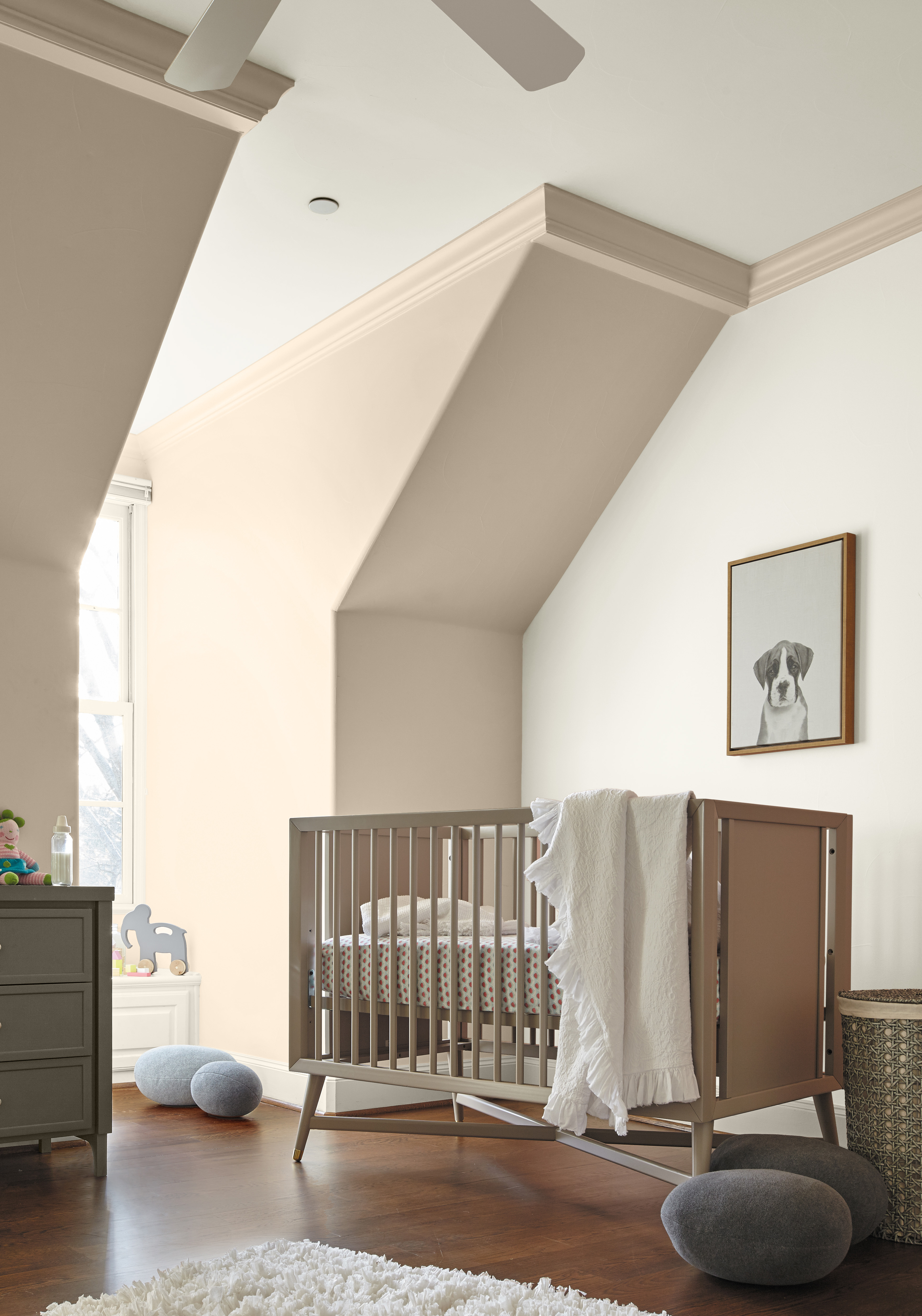 Cette image montre une chambre d’enfant avec des murs en avancée peints en beige clair, un lit de bébé blanc, une carpette blanche, des coussins épars sur un plancher en bois franc et un tableau représentant un chien au-dessus du lit.