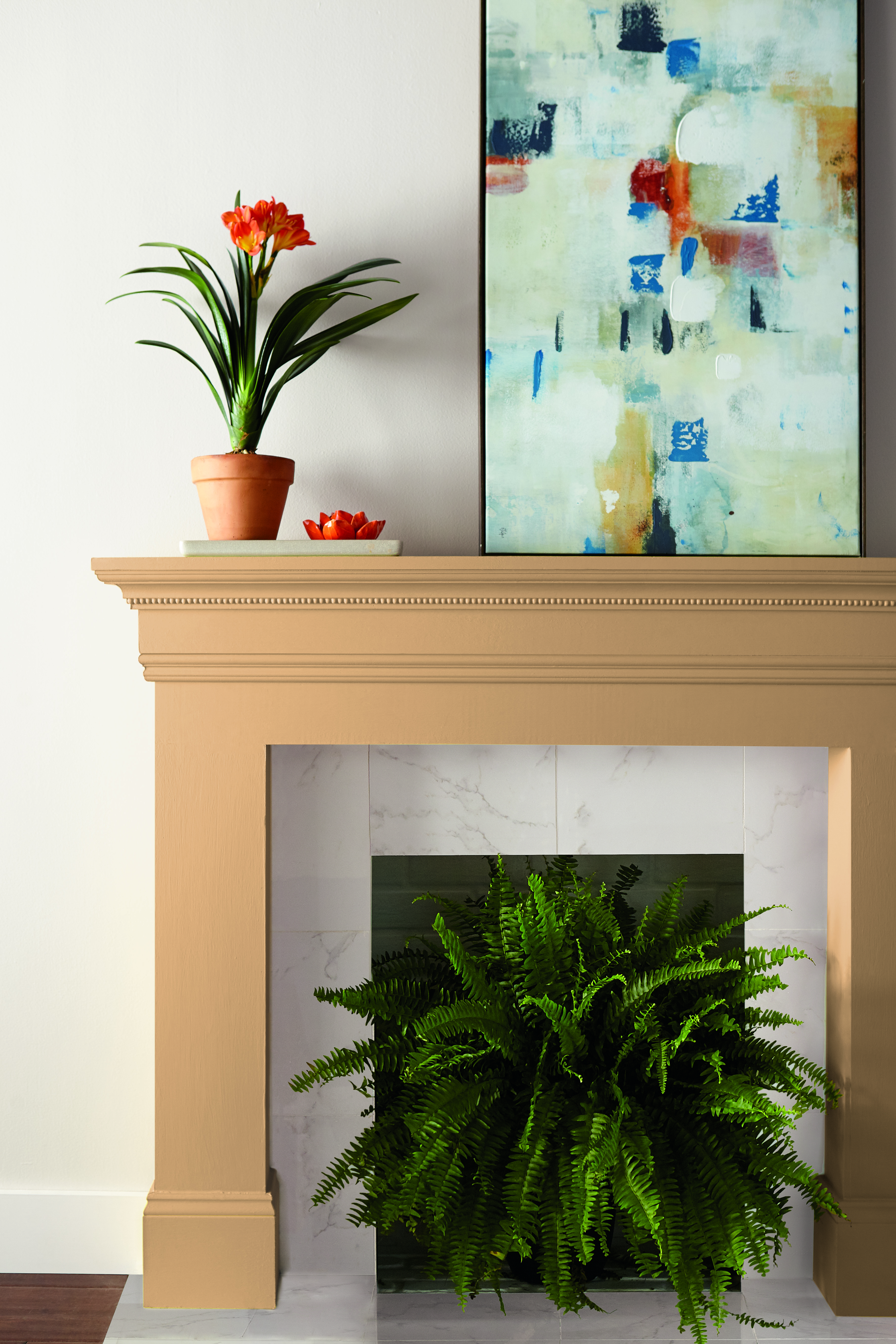 Cette image montre la façade d’un foyer peinte en jaune doré. Un pot de fleurs orange et une œuvre d’art abstraite sur le manteau de la cheminée, ainsi qu’une fougère verte à l’intérieur de l’âtre, parachèvent le décor sur fond de mur blanc.