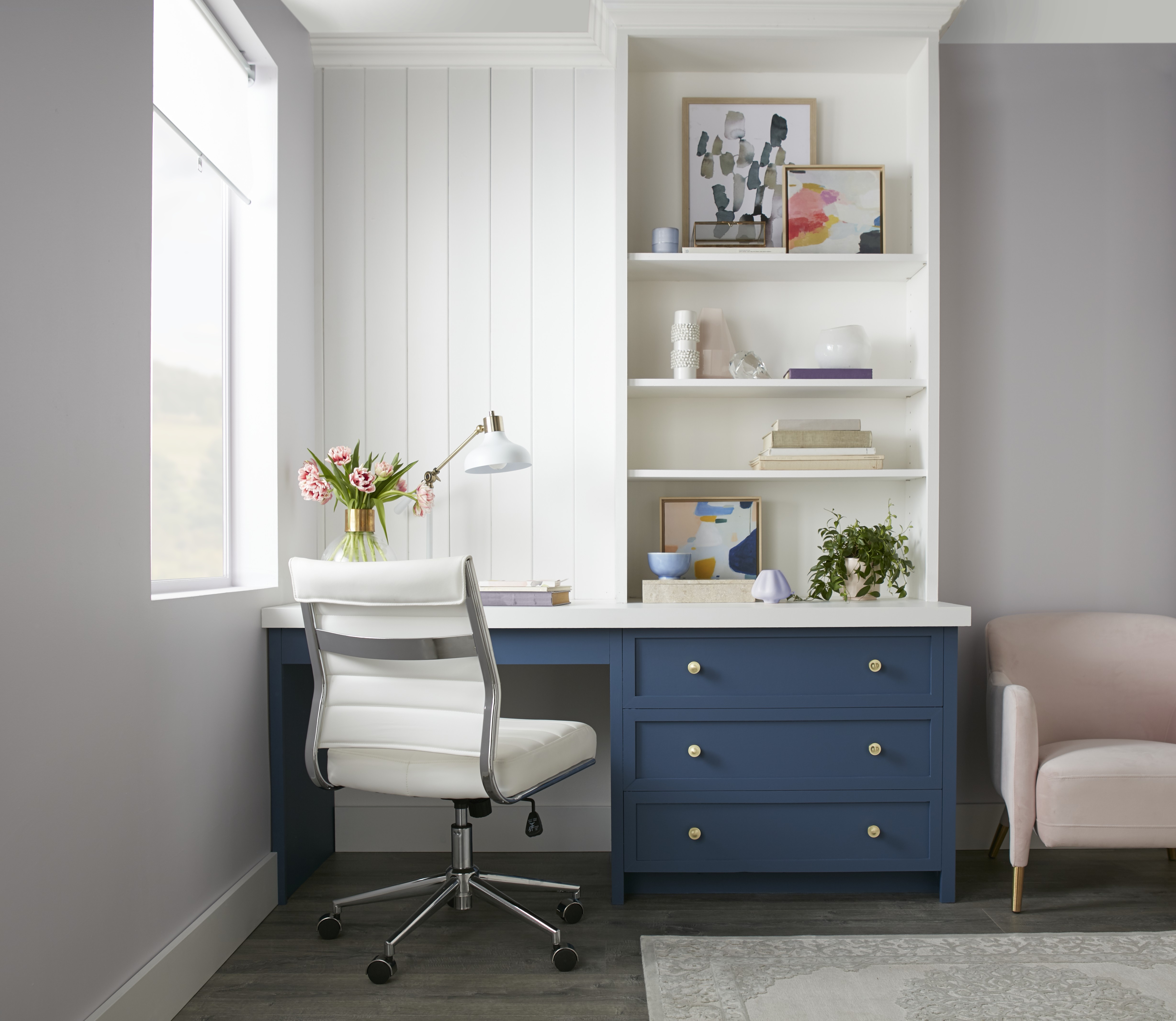 Cette image montre un espace de travail à domicile avec des murs peints en blanc, un bureau doté de tiroirs bleu marine et d’une surface de travail blanche, une chaise à roulettes également blanche, ainsi qu’un fauteuil de couleur lilas à côté du bureau.