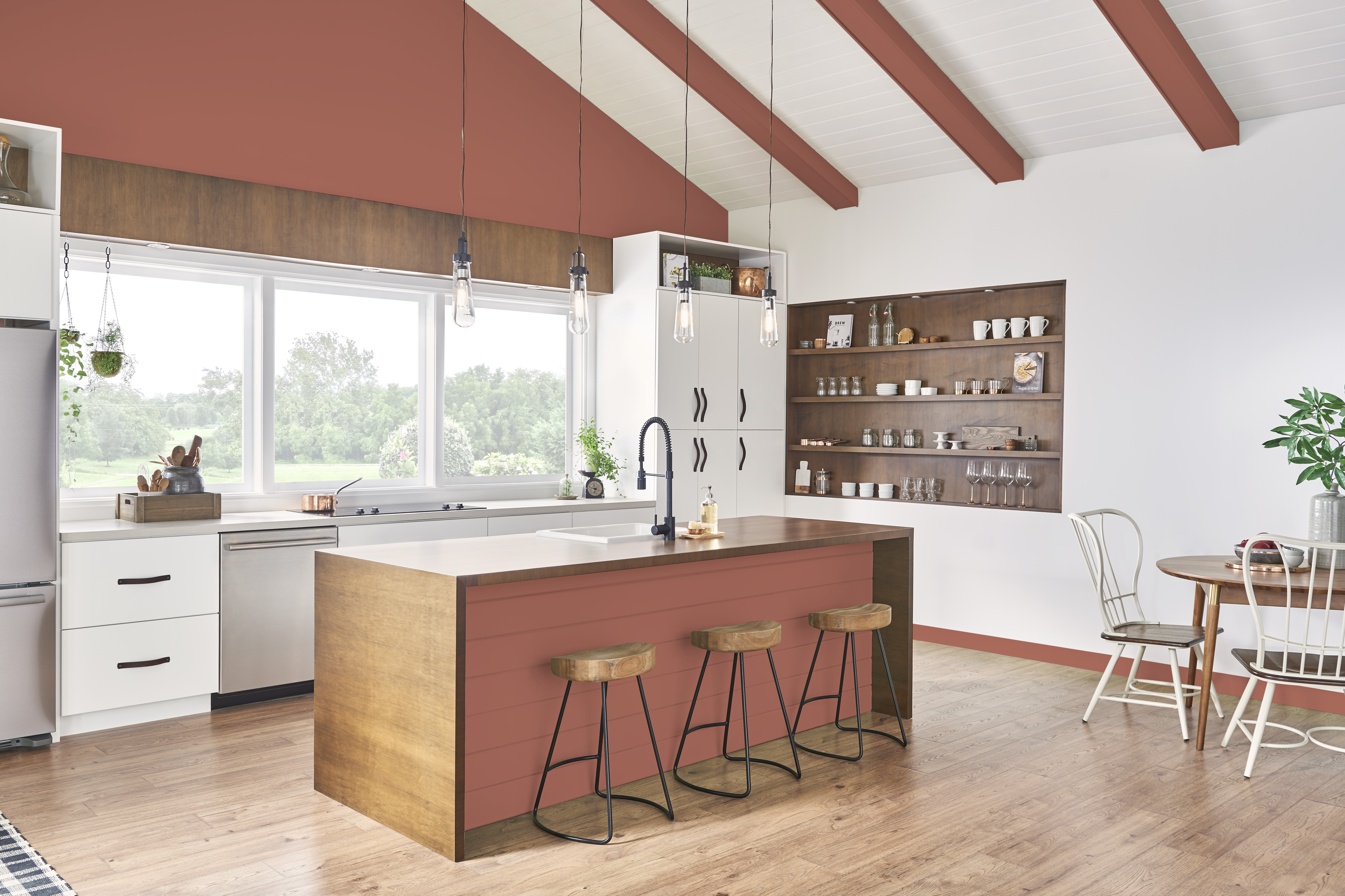 Cette image montre une cuisine moderne avec des murs blancs, l’un fenêtré et l’autre comportant des tablettes encastrées sans portes, ainsi que des poutres apparentes et la façade d’un îlot central peintes en terre cuite avec trois tabourets en bois.
