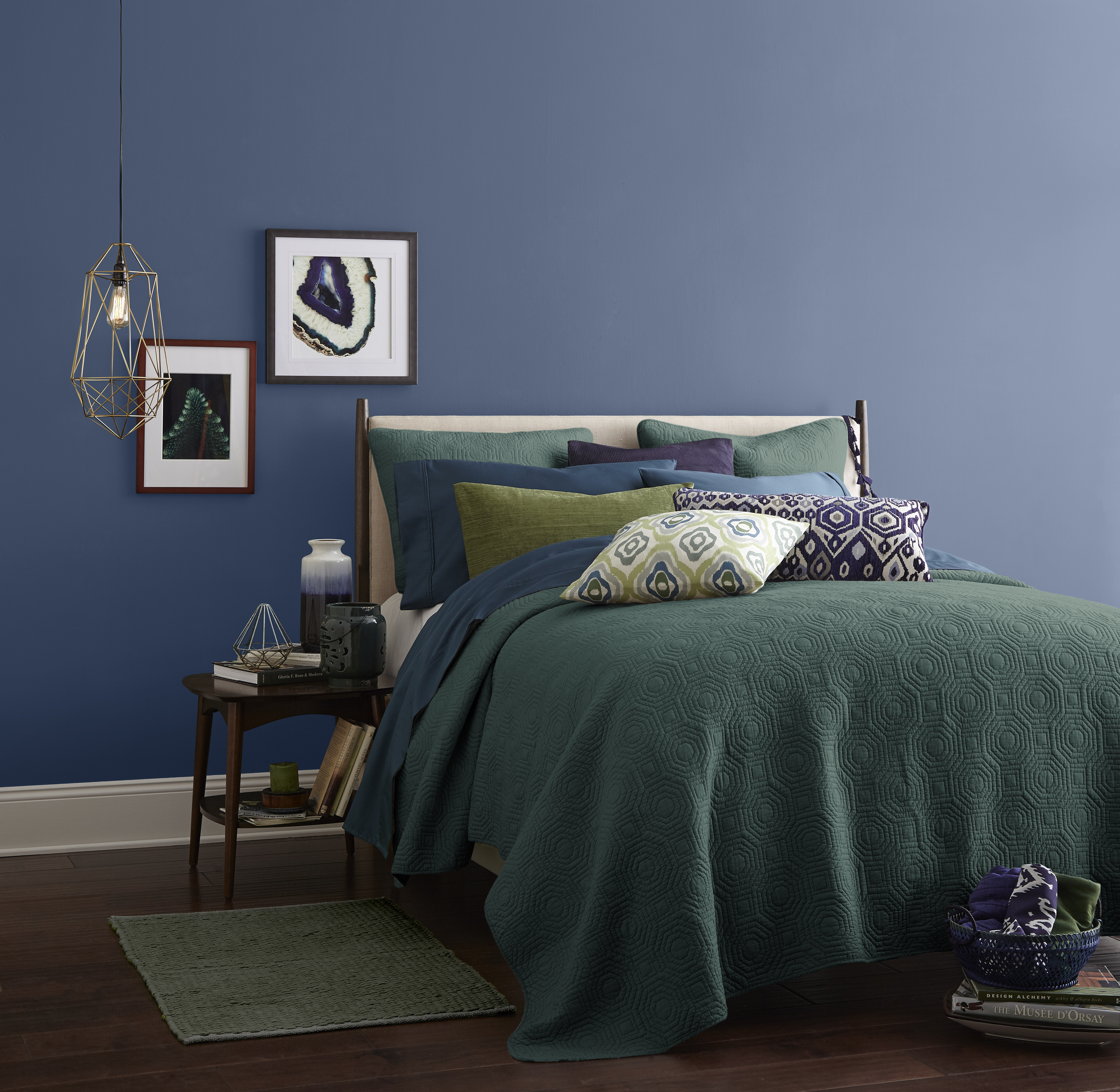 Cette image montre une chambre à coucher dont le mur est peint dans une teinte bleue apaisante. La literie aux couleurs de verroterie se décline dans des tons de vert, de bleu et de violet.