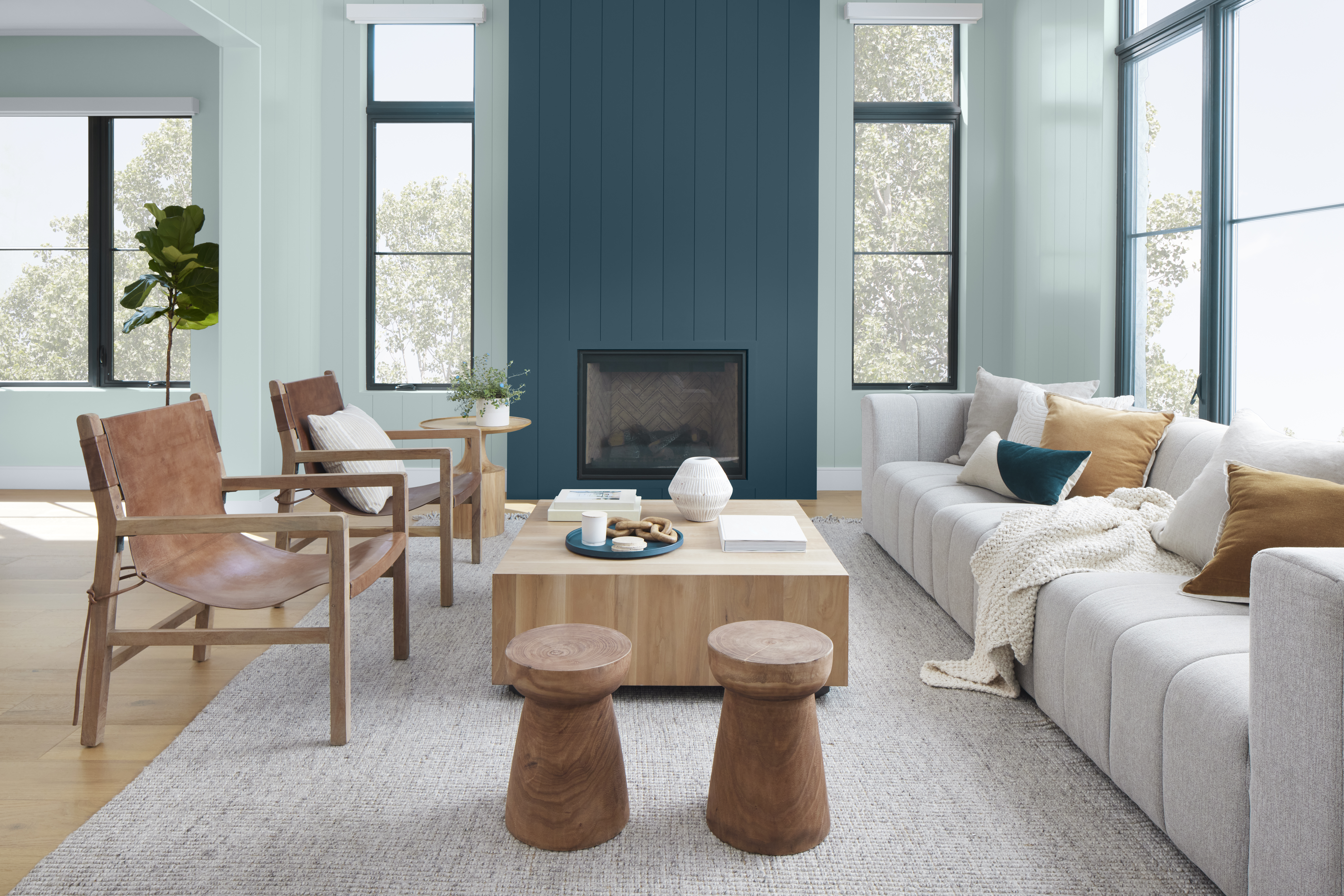 Cette image montre un salon moderne baigné de lumière naturelle et dont les murs peints en Aqueux font contraste avec le mur de la cheminée à parement à feuillures peint en un bleu profond appelé Bleu nocturne. L’espace comprend un long canapé gris clair, deux confortables fauteuils en bois, des coussins blancs et fauves ainsi qu’une table basse carrée en bois massif ornée d’objets décoratifs.