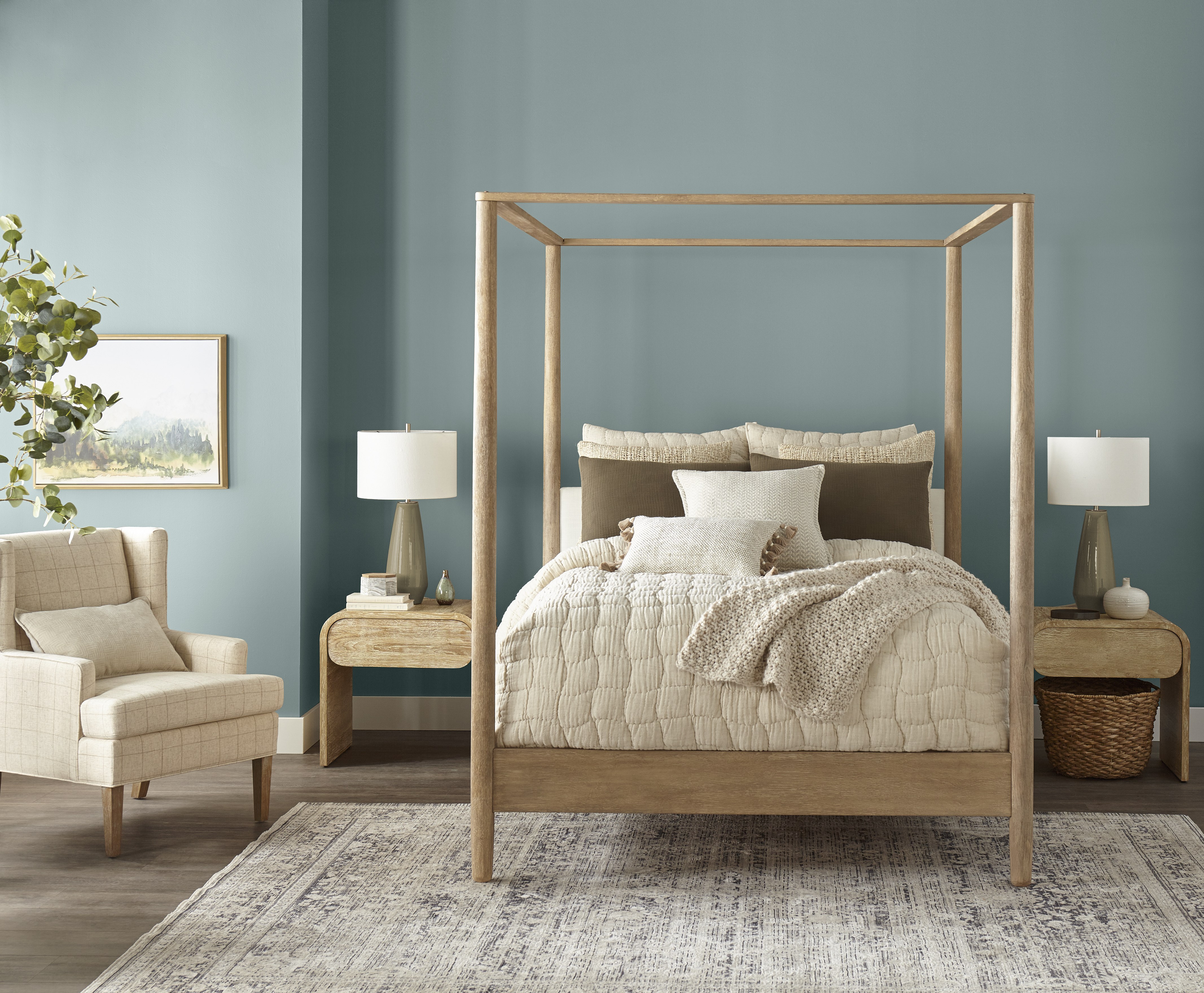 Cette image montre une chambre à coucher douillette avec un lit à baldaquin en bois et une literie de couleur neutre où se superposent une couette matelassée et un jeté de lit texturé. Les murs peints en un bleu clair doux créent une ambiance calme et équilibrée. Le lit est flanqué de deux tables de chevet en bois sur chacune desquelles repose une lampe à abat-jour circulaire. La pièce comporte également un fauteuil rembourré de couleur claire, un tapis au motif irrégulier et une plante verte en pot qui apporte une touche de verdure.