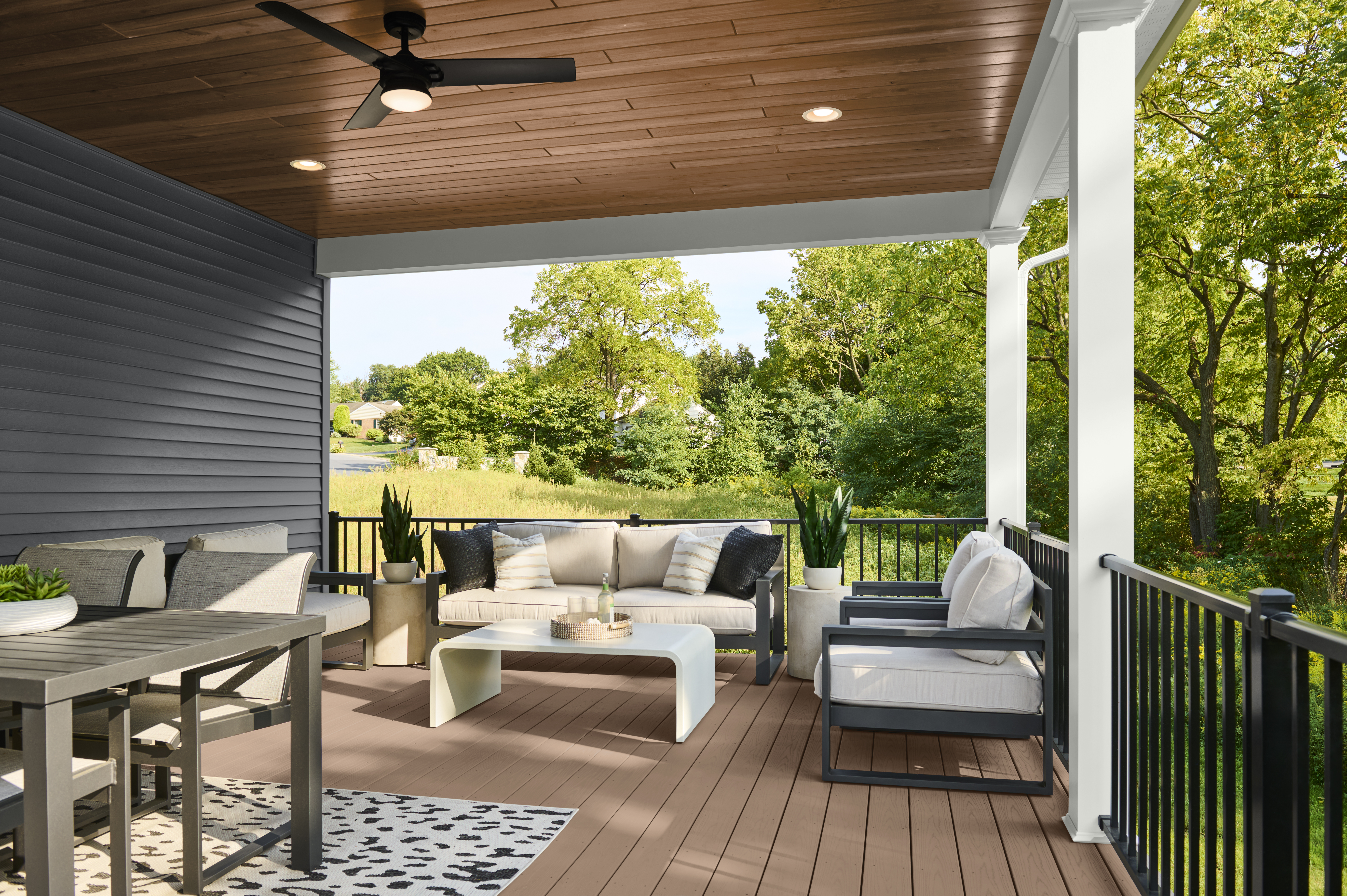 Cette image montre une véranda donnant sur un paysage verdoyant. Le plafond de la véranda est en bois teint. La terrasse en bois est teinte d’une couleur assortie et meublée de fauteuils d’extérieur, d’une table à manger et d’un ventilateur de plafond.