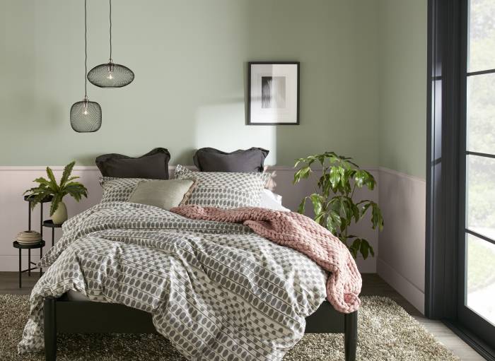 Cette image montre une chambre à coucher sereine, dont les murs rose pâle et vert sauge font contraste avec le lit aux montants sombres et habillé d’une épaisse literie à motifs blanc et gris. Les oreillers adossés aux coussins anthracite créent un air d’élégance avec le jeté rose poudré au pied du lit. Deux petites lampes suspendues éclairent un côté du lit, tandis qu’une œuvre en noir et blanc du côté droit apporte une touche artistique à l’ensemble.
