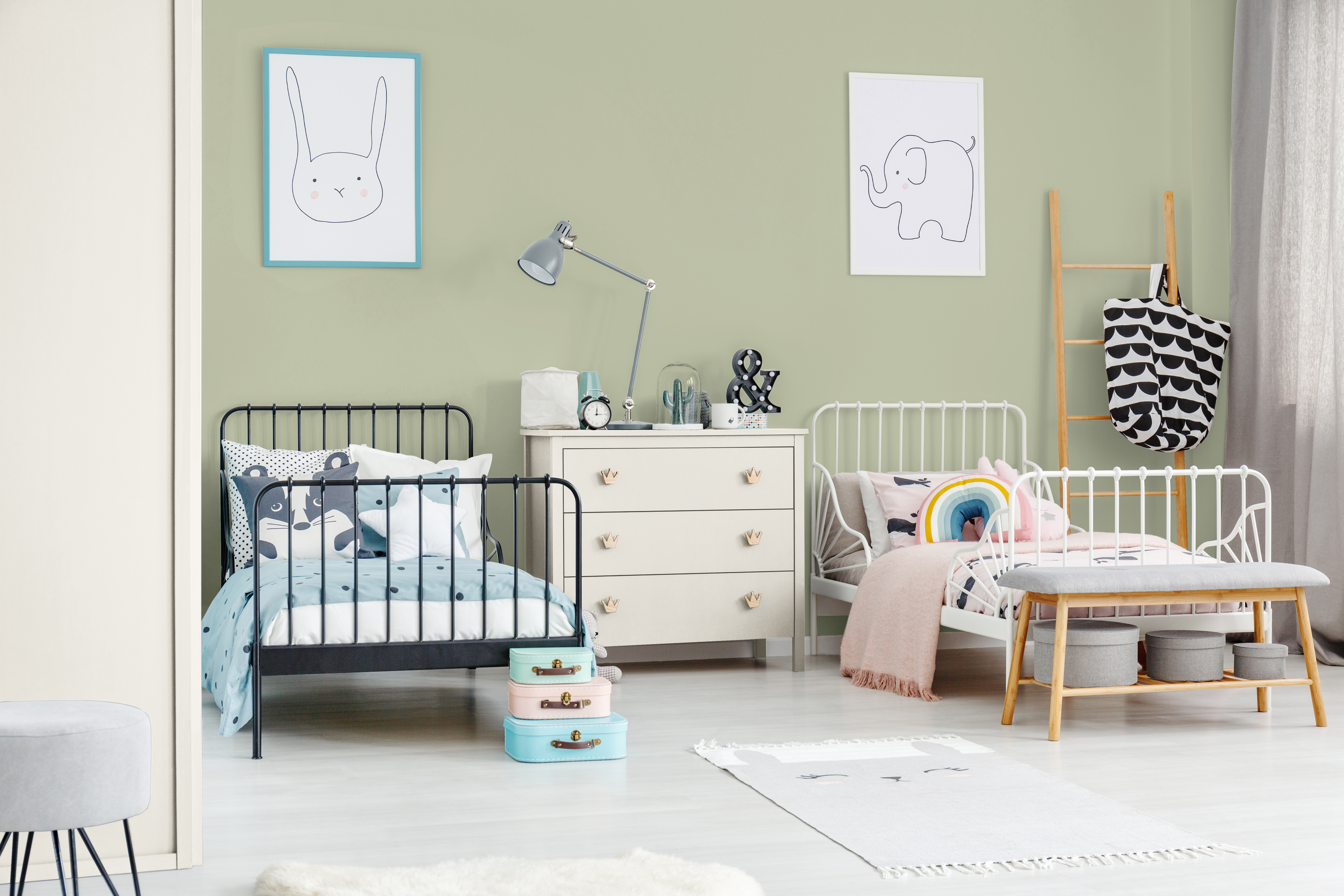 Cette image montre une chambre d’enfants partagée avec un mur d’accent vert tendre, deux lits jumeaux avec tête et pied en barreaux de métal (l’un noir et l’autre blanc), des articles de mobilier en bois clair, une literie blanche et bleu ciel d’un côté, et blanche et rose de l’autre, ainsi que des dessins d’animaux ludiques au-dessus des lits.