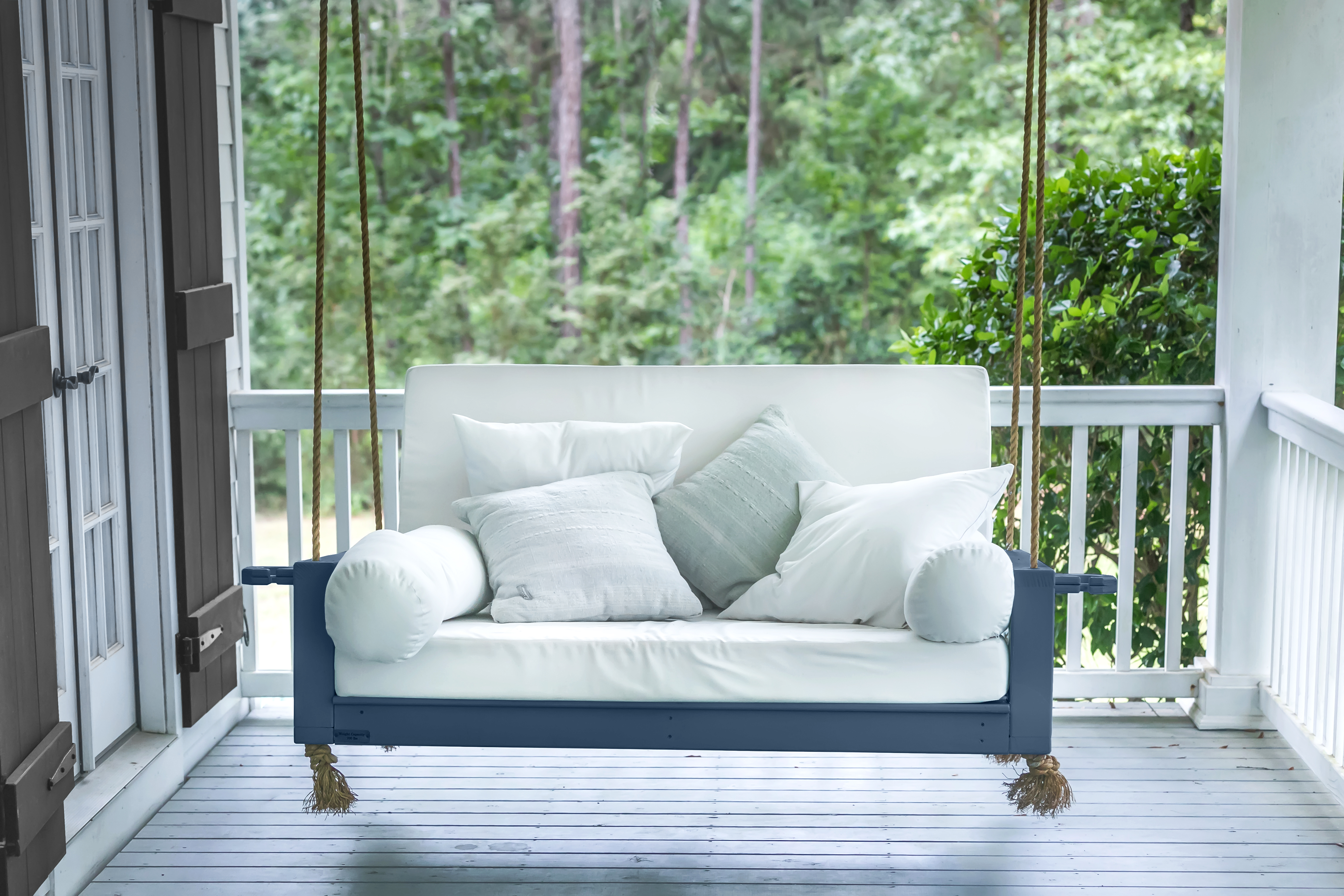 Alt text: Cette image montre une balancelle peinte en bleu marine qui contraste avec la blancheur des coussins du siège et du dossier, eux-mêmes agrémentés de petits coussins décoratifs aux tons pâles.