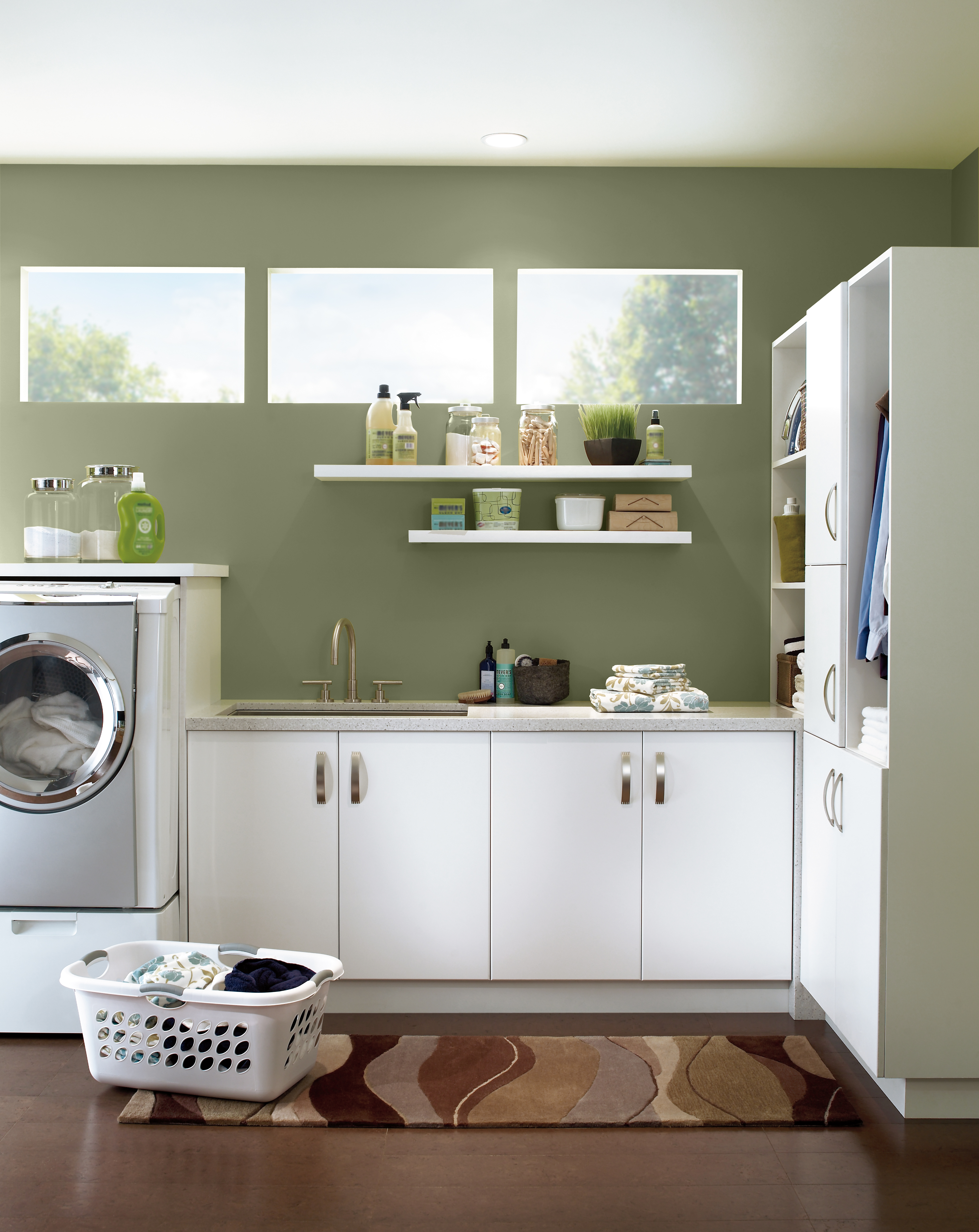 Cette image montre une salle de lavage verte avec des murs en teinte «Écologique». On aperçoit une série d’armoires blanches, deux tablettes blanches, une laveuse à chargement frontal et un panier à lessive bien rempli, le tout créant un style épuré mais néanmoins fonctionnel.