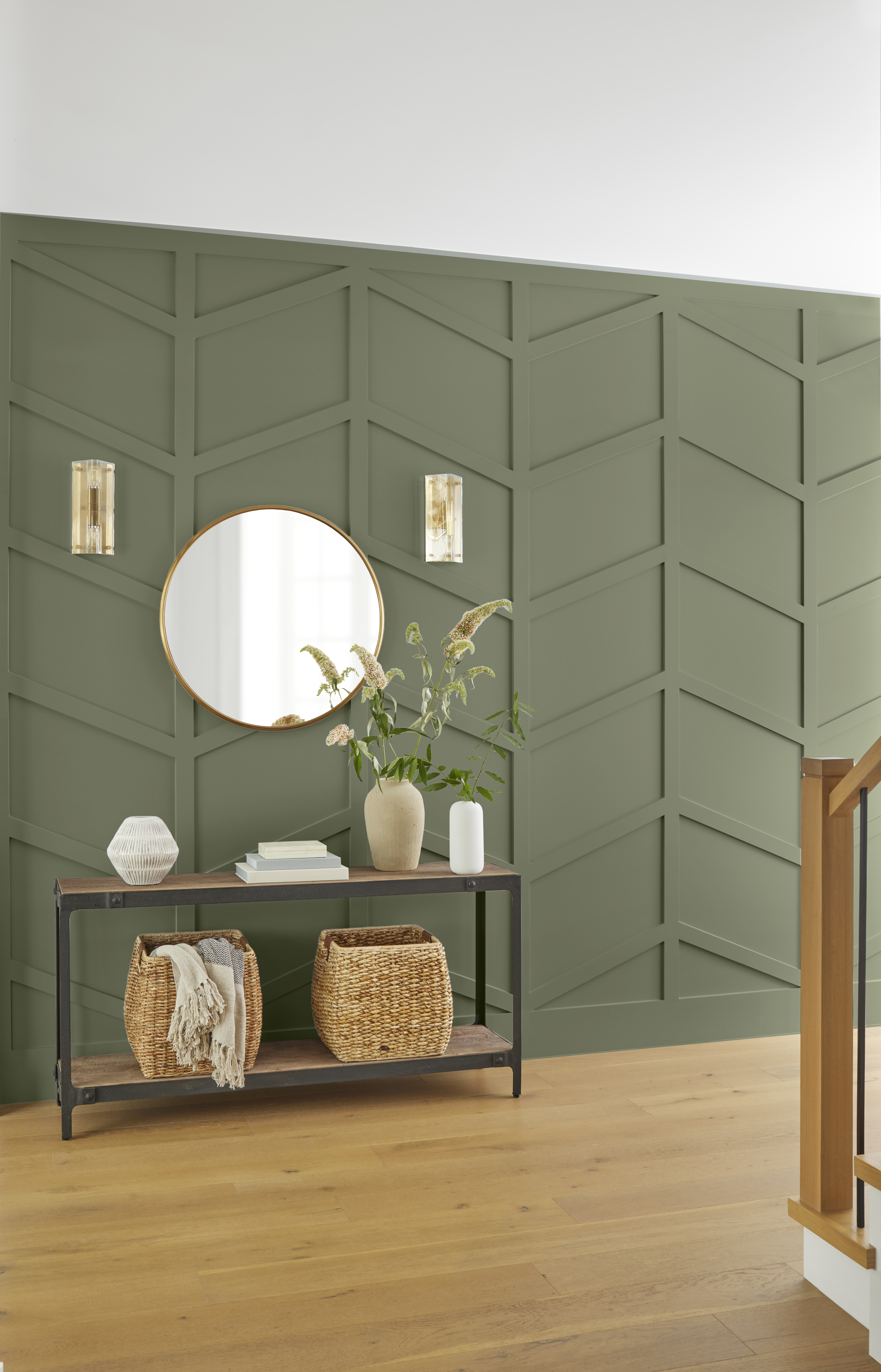 Cette image montre un hall d’entrée avec un mur lambrissé en chevrons et peint en teinte «Écologique», auquel sont accrochés un miroir rond et deux appliques murales. On y voit également un meuble console où se côtoient deux paniers d’osier sur la tablette du bas et quelques objets décoratifs sur celle du haut.