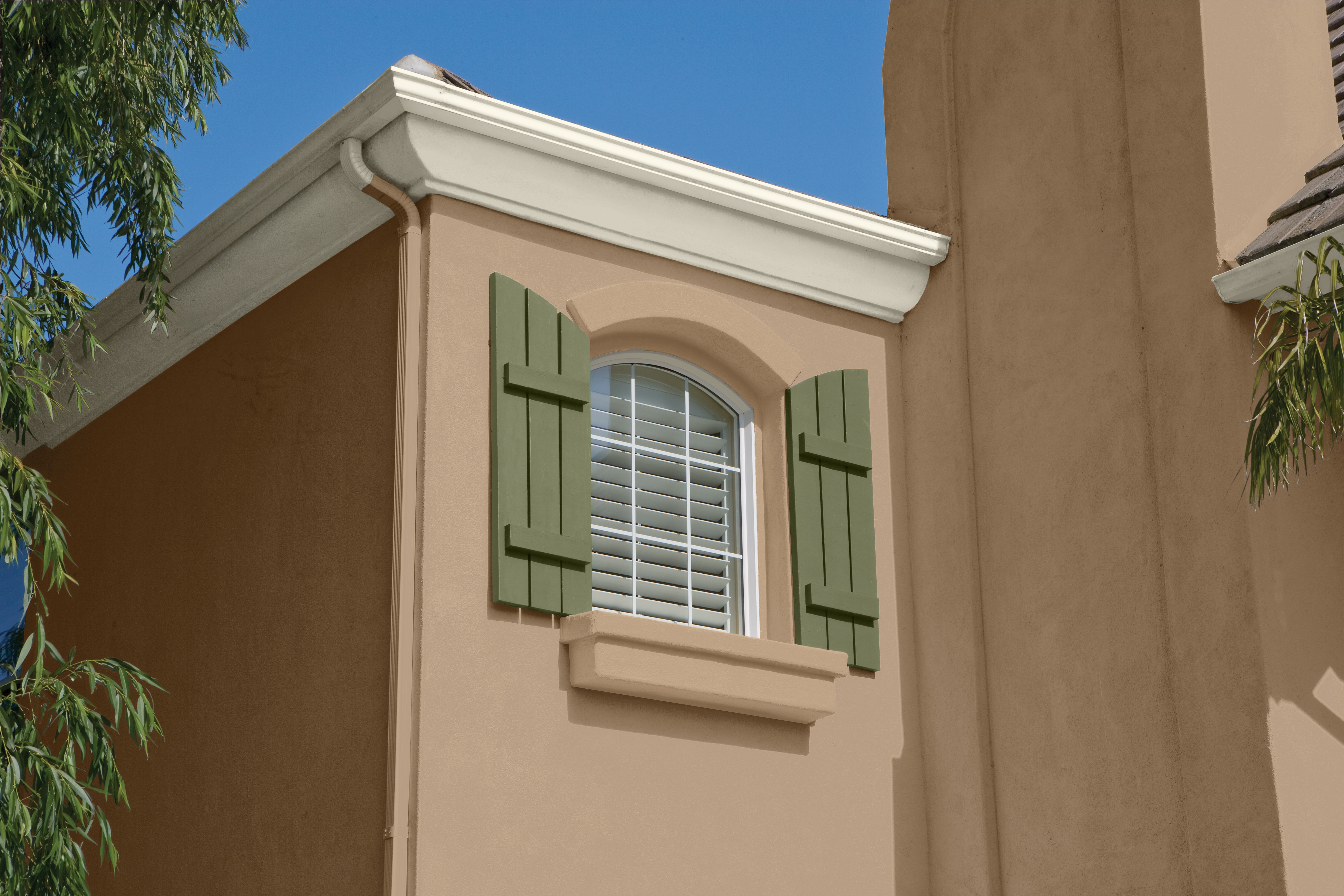 Cette image montre l’aile extérieure d’une maison revêtue d’un stucco de couleur beige, où l’on aperçoit une fenêtre en arche parée d’un store vénitien blanc et de volets verts décoratifs.