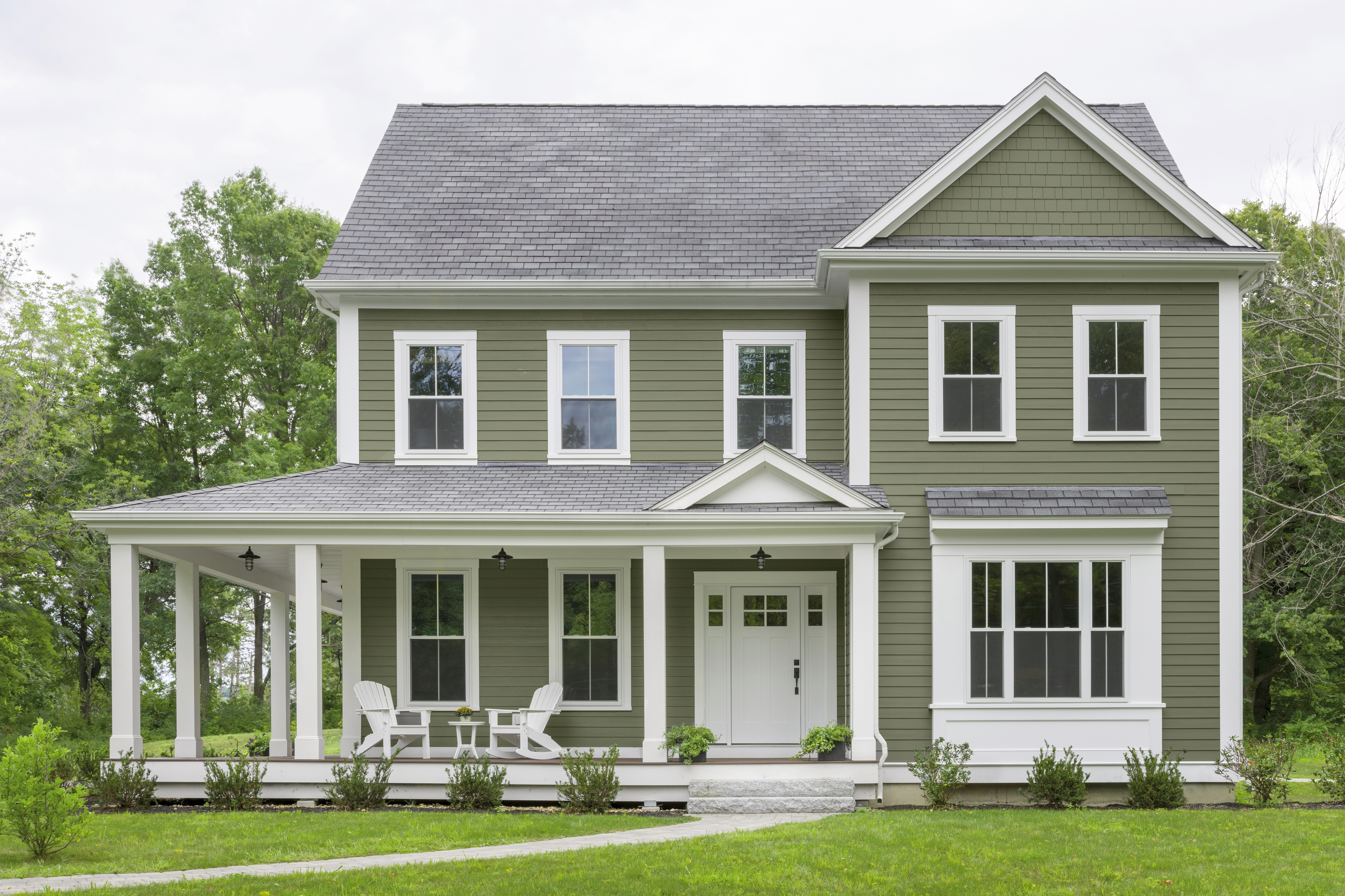 Cette image montre une maison à deux étages de style maison de ferme. Elle comporte un bardage peint en vert, des garnitures blanches, une galerie enveloppante et une grande pelouse verte.