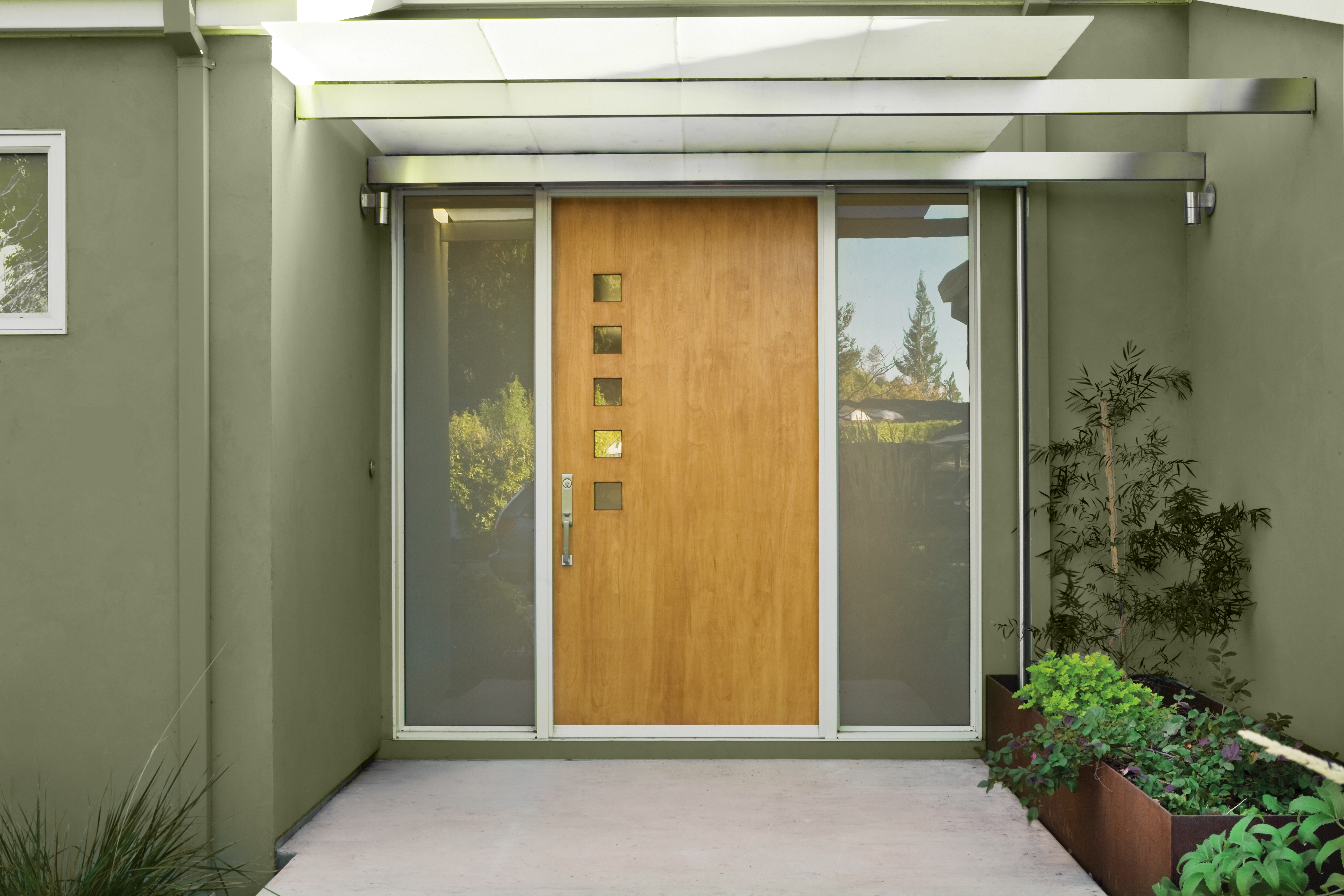 Cette image montre une entrée de maison de style moderne avec une porte en bois blond, des panneaux de vitre givrés de chaque côté, un auvent métallique et des jardinières. Le corps de la maison est revêtu d’un stucco peint en vert.