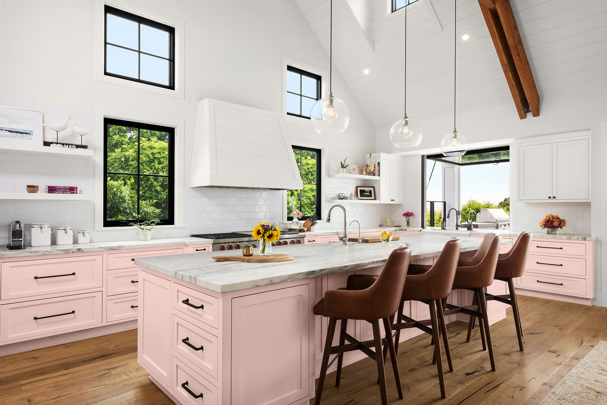 Cette image montre une cuisine dominée par la couleur rose, avec des armoires et tiroirs peints en rose pastel, des comptoirs en marbre, des fenêtres au cadrage noir, des luminaires suspendus et un vaste îlot avec des chaises de bar en cuir brun.