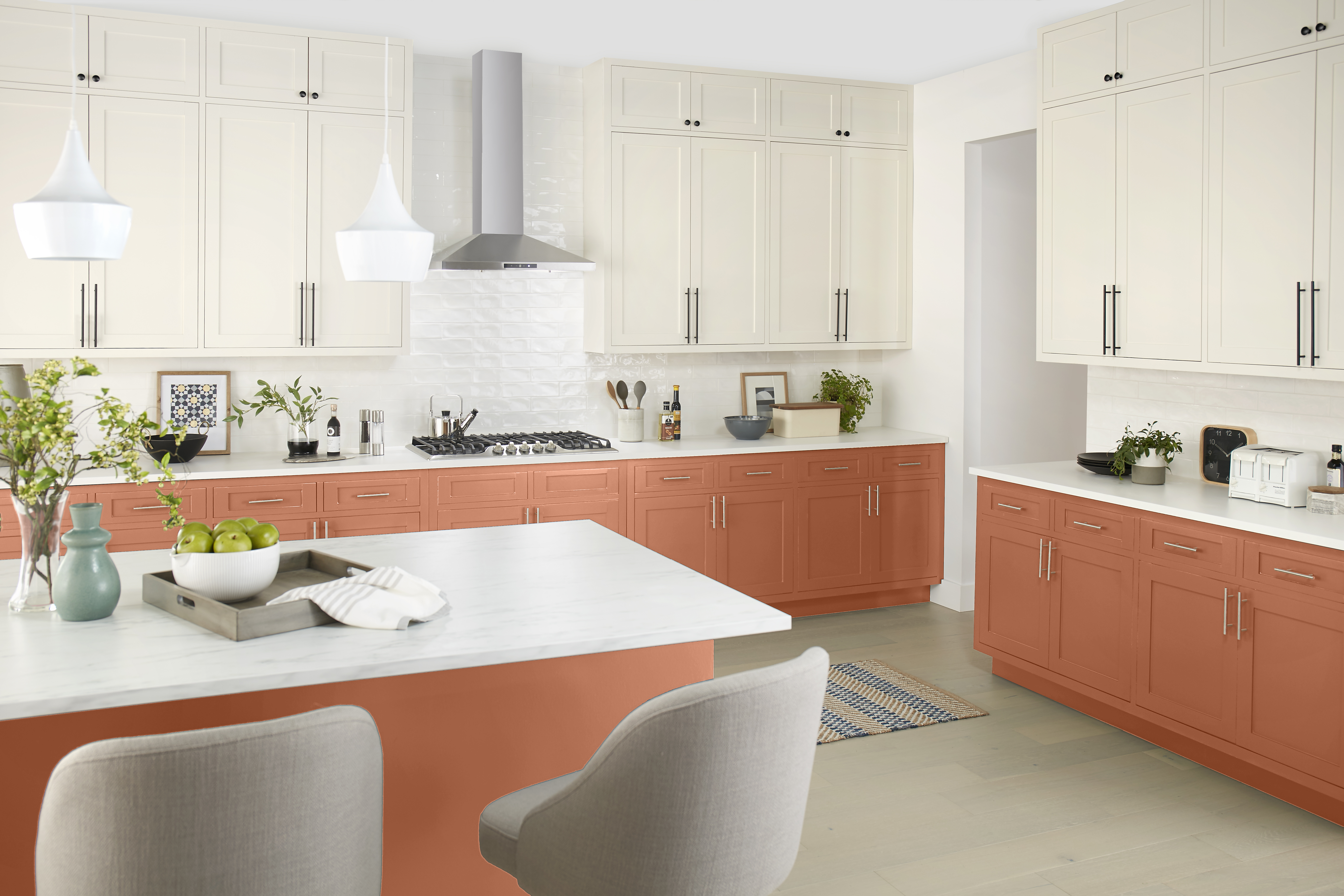 Cette image montre une cuisine bien éclairée avec des armoires du bas peintes en terre cuite, des armoires du haut de couleur crème, des comptoirs blancs et des luminaires suspendus de style moderne.