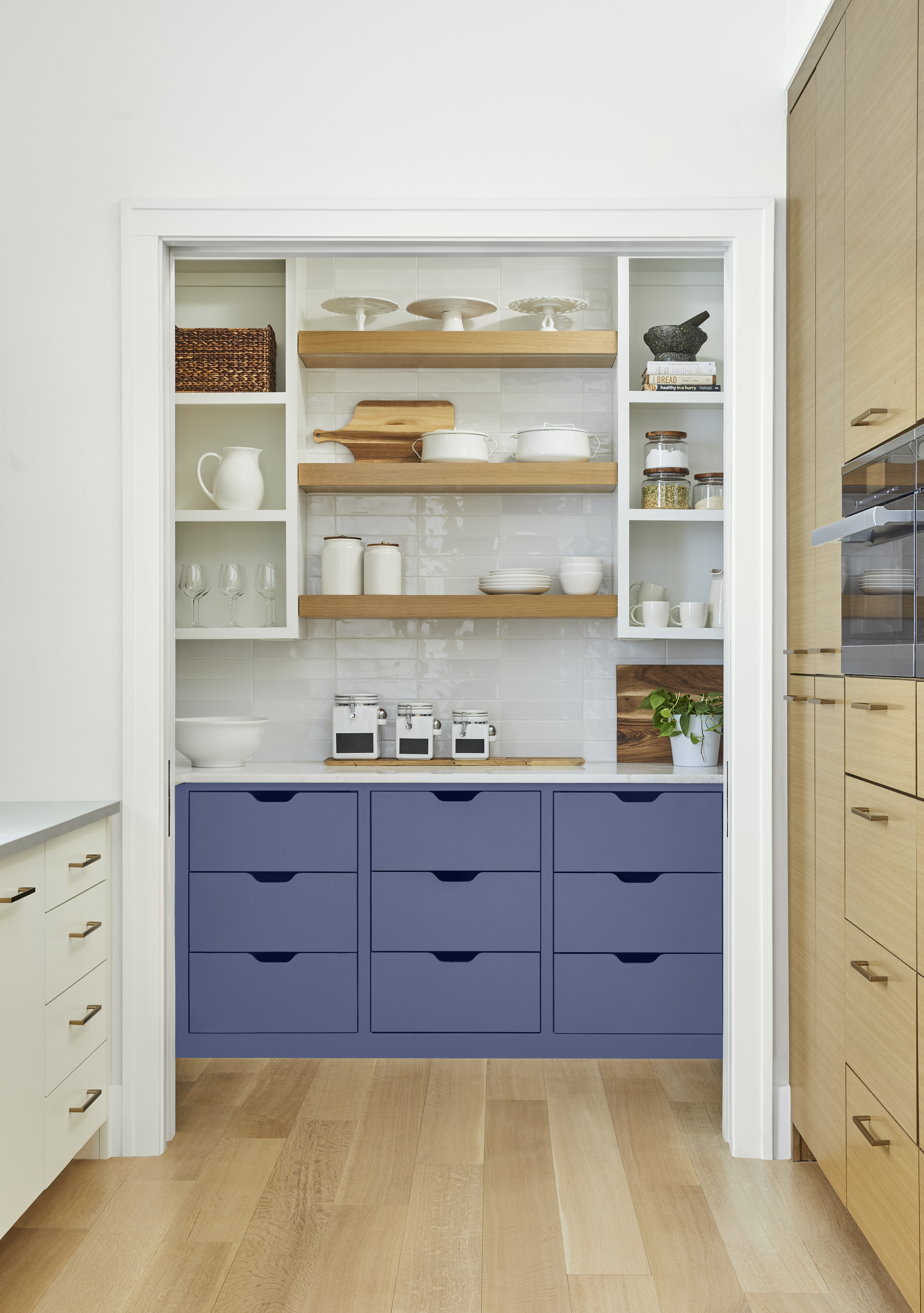 Cette image montre un coin garde-manger dont les tiroirs peints en bleu-violet apportent un éclat de couleur inattendu à un espace à vocation purement fonctionnelle. Sur le mur du fond, on aperçoit trois tablettes en bois encastrées entre deux armoires blanches sans portes. Un parquet en bois blond, flanqué d’un meuble de rangement blanc d’un côté et d’armoires en bois naturel de l’autre, mène au garde-manger.