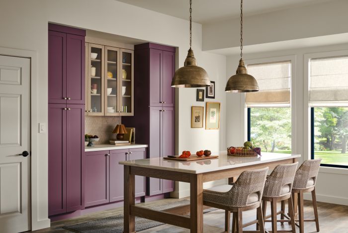 Cette image montre un coin repas riche et invitant avec des armoires rouge-violet, une table en bois, des chaises en rotin, deux luminaires suspendus en laiton et deux grandes fenêtres avec vue sur des arbres.