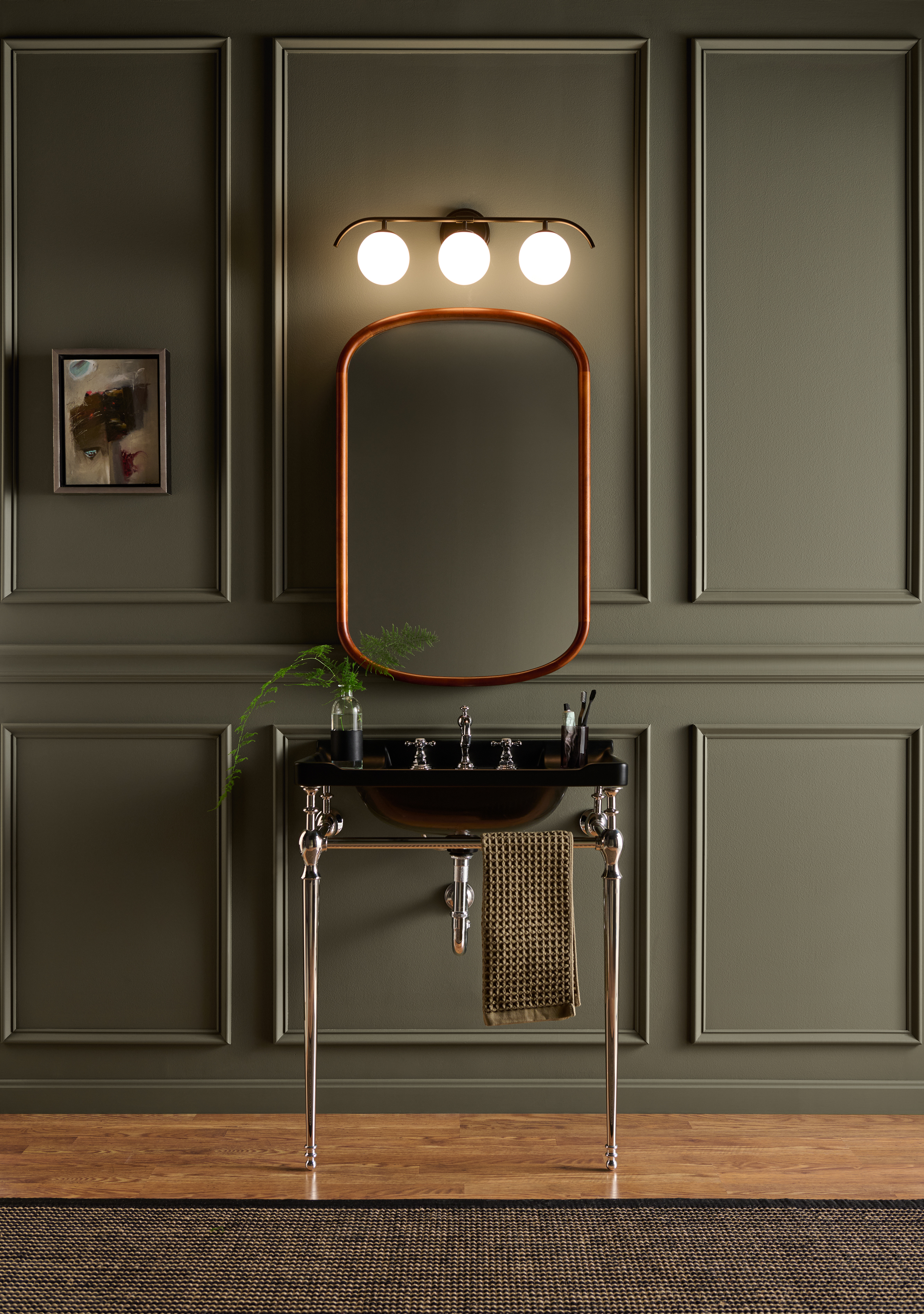 Cette image montre une élégante salle de bain minimaliste agrémentée d’un mur lambrissé peint en teinte Terre à terre de BEHR, d’un lavabo noir soutenu par un montant chromé, d’un miroir aux contours arrondis, d’un jeu de lumière sur rail au-dessus du lavabo et d’un parquet de bois au ton chaud.